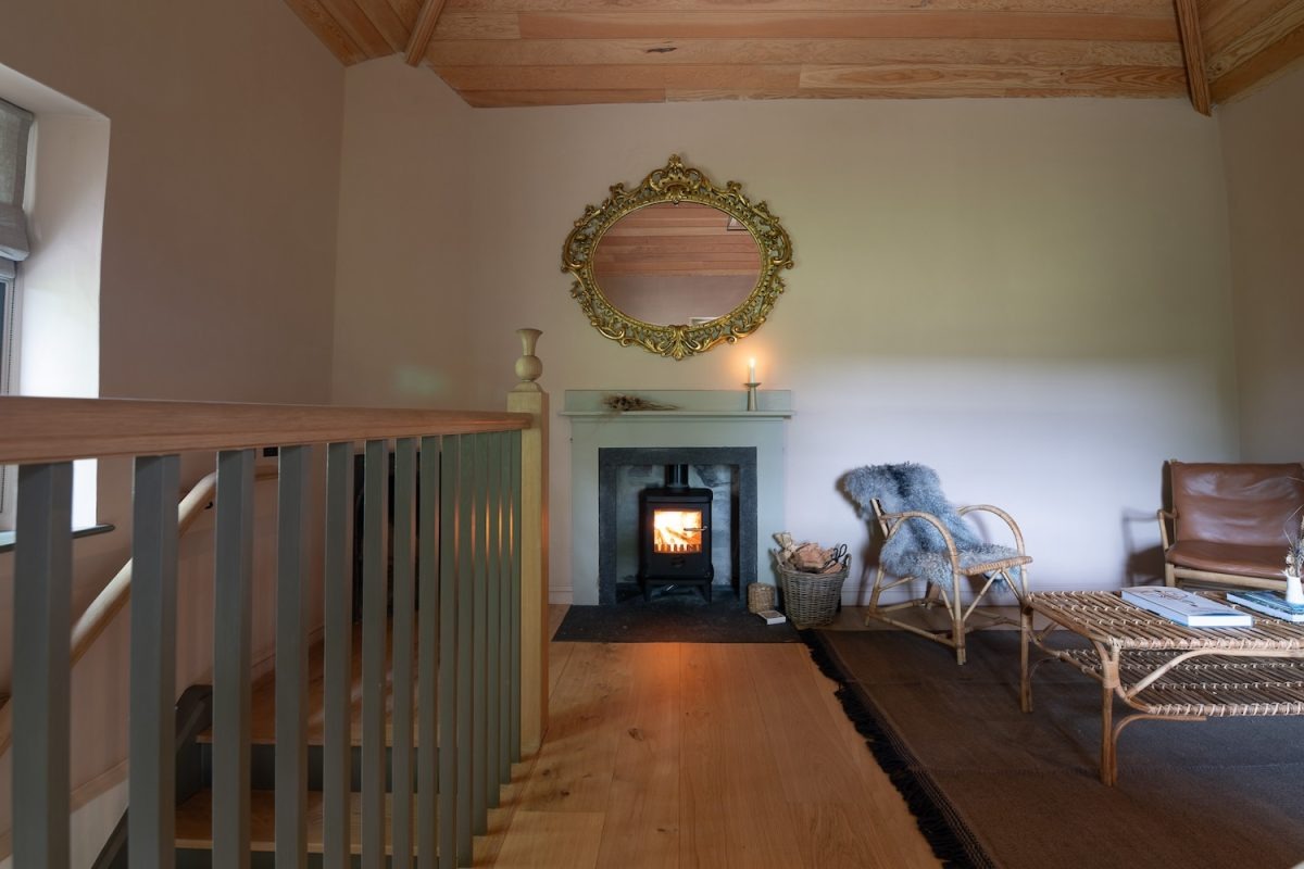 A wood burning stove is alight beneath a gilt-framed oval mirror, a wicker chair with a sheepskin throw and a low table completing the mezzanine sitting area.