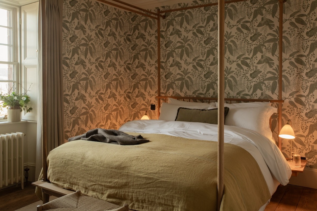 A four-poster bed with an olive throw stands against patterned wallpaper, a wicker armchair and small stool at its foot.