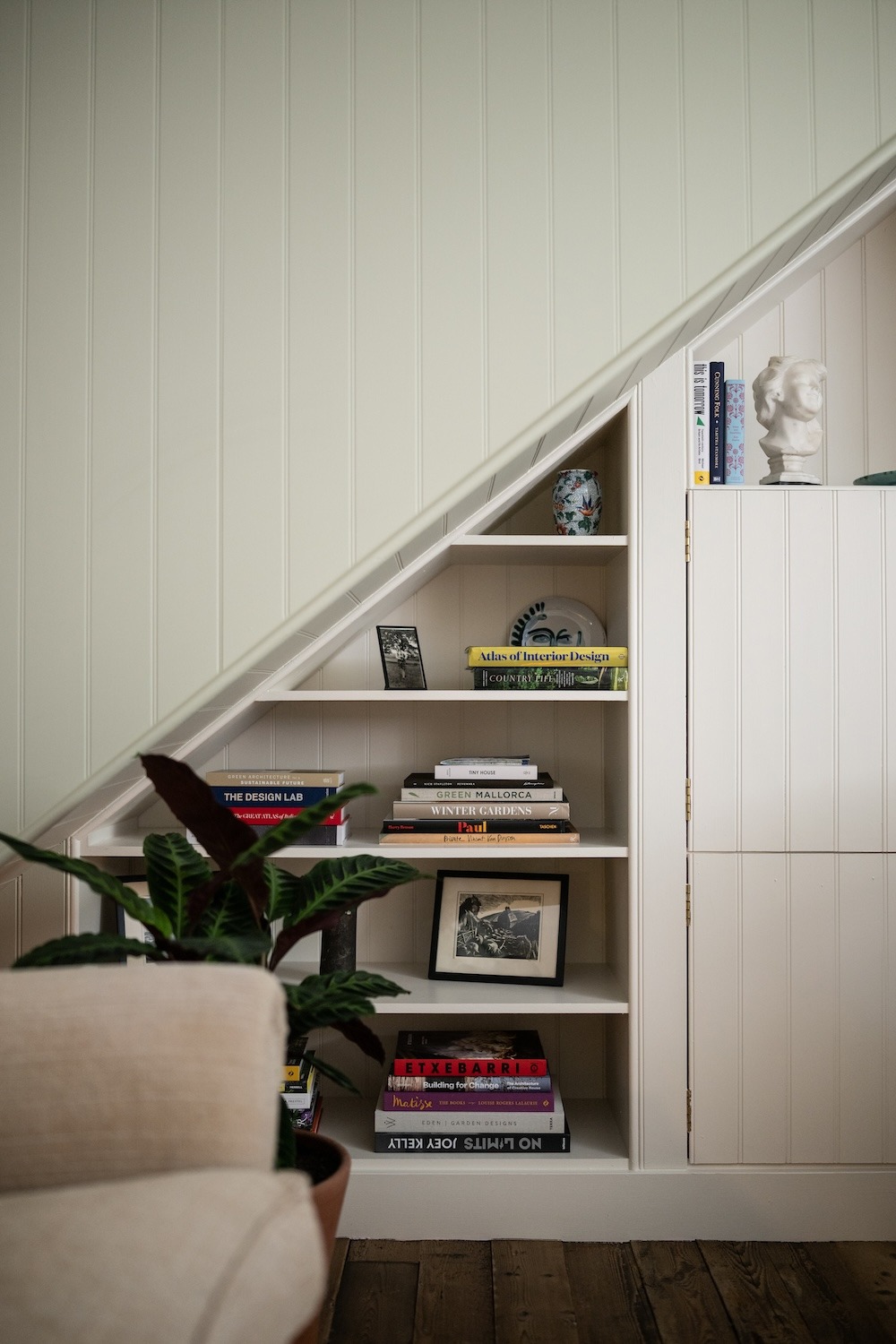 Books including an "Atlas of Interior Design" are stacked on shelves built into a sloped alcove, a potted plant and framed photograph among them.