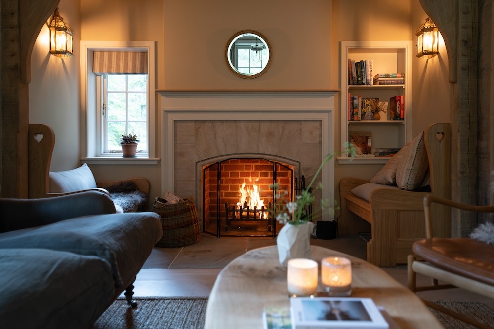 A wood burning stove burns behind a mesh guard in a brick fireplace, a round mirror above the mantel and bookshelves either side.