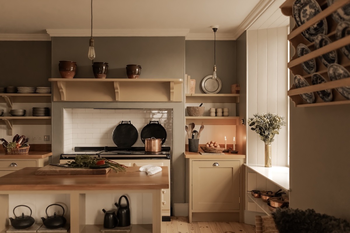 A kitchen island with a bowl of vegetables stands before a range cooker set into a tiled surround, open shelving of crockery either side.
