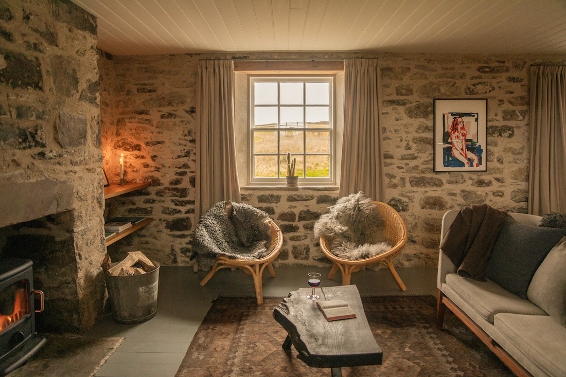 Two wicker chairs with sheepskin throws sit before a window, a sofa and low table with a book and wine glass in the foreground of a stone-walled room.
