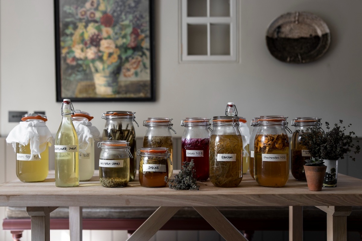 Jars of preserves and infused vinegars with handwritten labels are lined up on a wooden table before a floral still life painting.