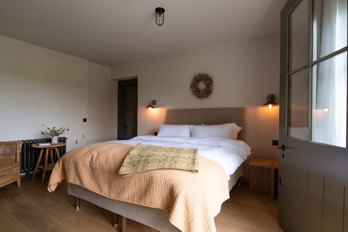 A bed with a peach throw and a checked cushion sits below a woven wreath, wall lamps lit either side and a stable door open to the right.