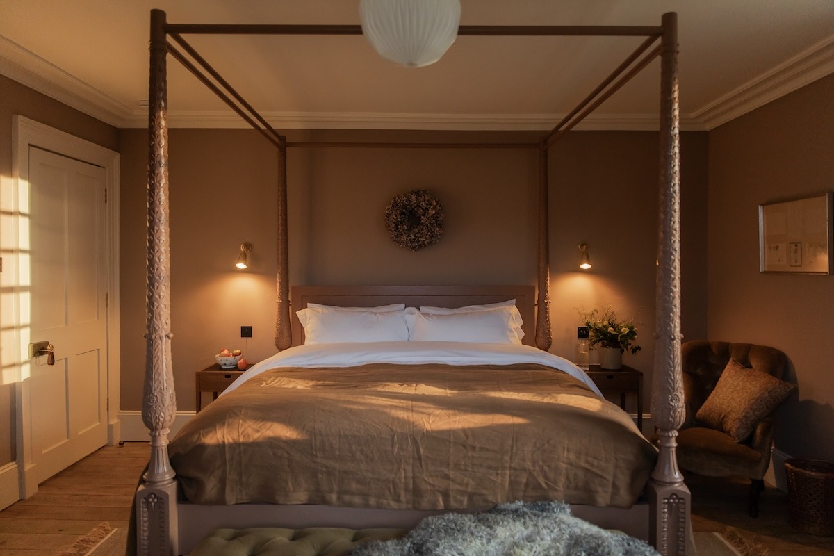 A four-poster bed with taupe bedding stands beneath a wreath hung on the wall, wall sconces lit either side and a wicker chair in the corner.