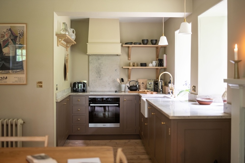 A kitchen with brown cabinetry and a marble splashback, open shelving of crockery beside a cooker and hood, a framed print on the wall to the left.