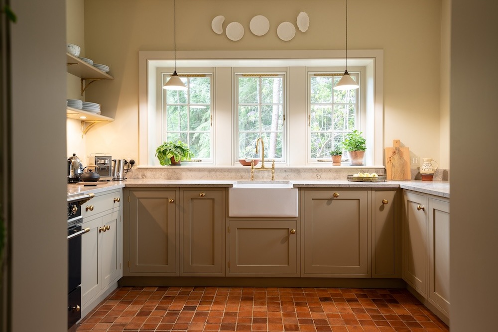 A U-shaped kitchen with sage green units and a belfast sink stands below a row of windows, decorative plates displayed above.