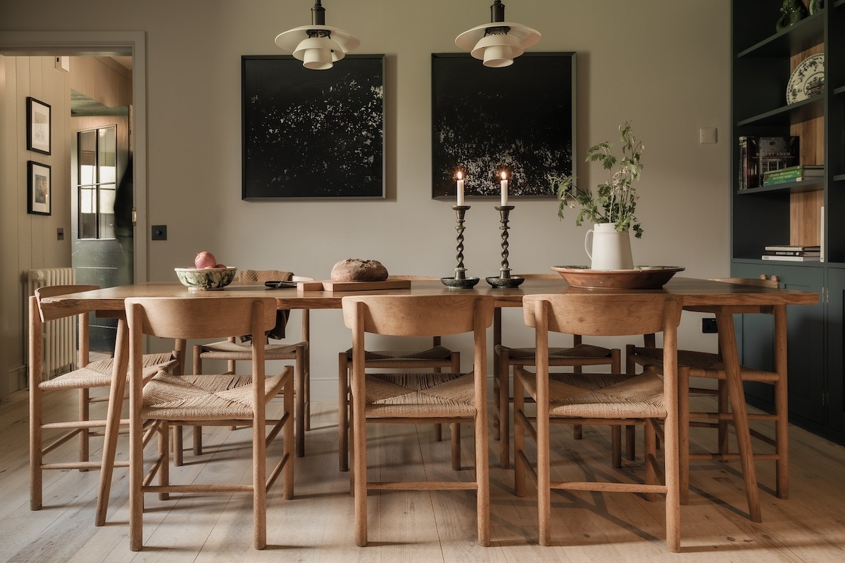 A long wooden dining table set with fruit and candles stands before dark abstract prints, woven-seat chairs around it.