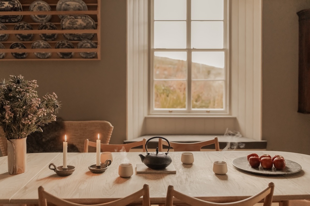 A table set with candles, teacups and a bowl of tomatoes stands before a window, decorative plates displayed on a shelf to the left.