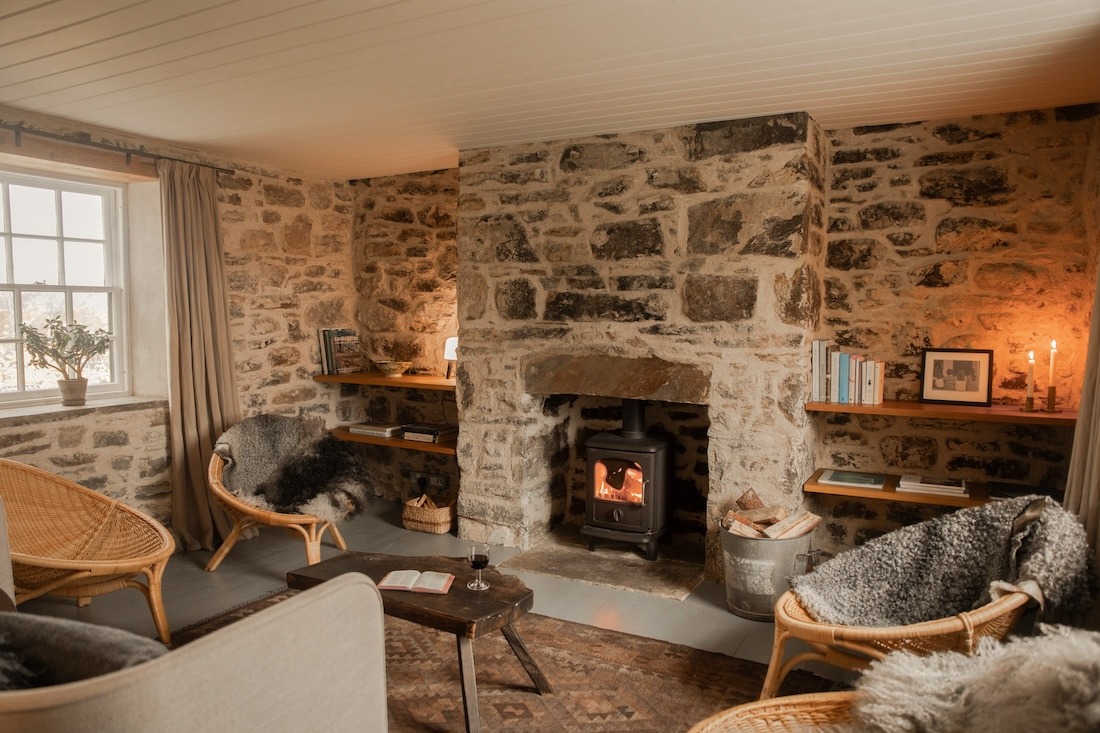 A wood burning stove burns in a stone fireplace between built-in shelving, wicker chairs draped with sheepskins on either side.