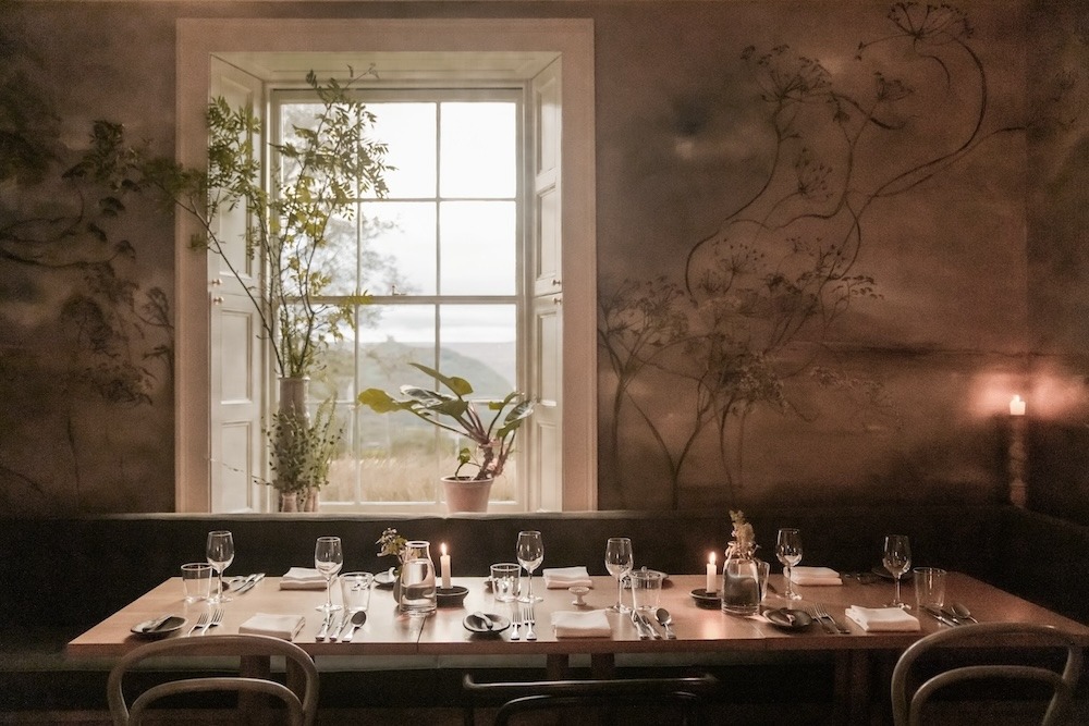 A long dining table set for a formal meal with candles and glasses stands before a tall window, hand-painted branches decorating the walls.