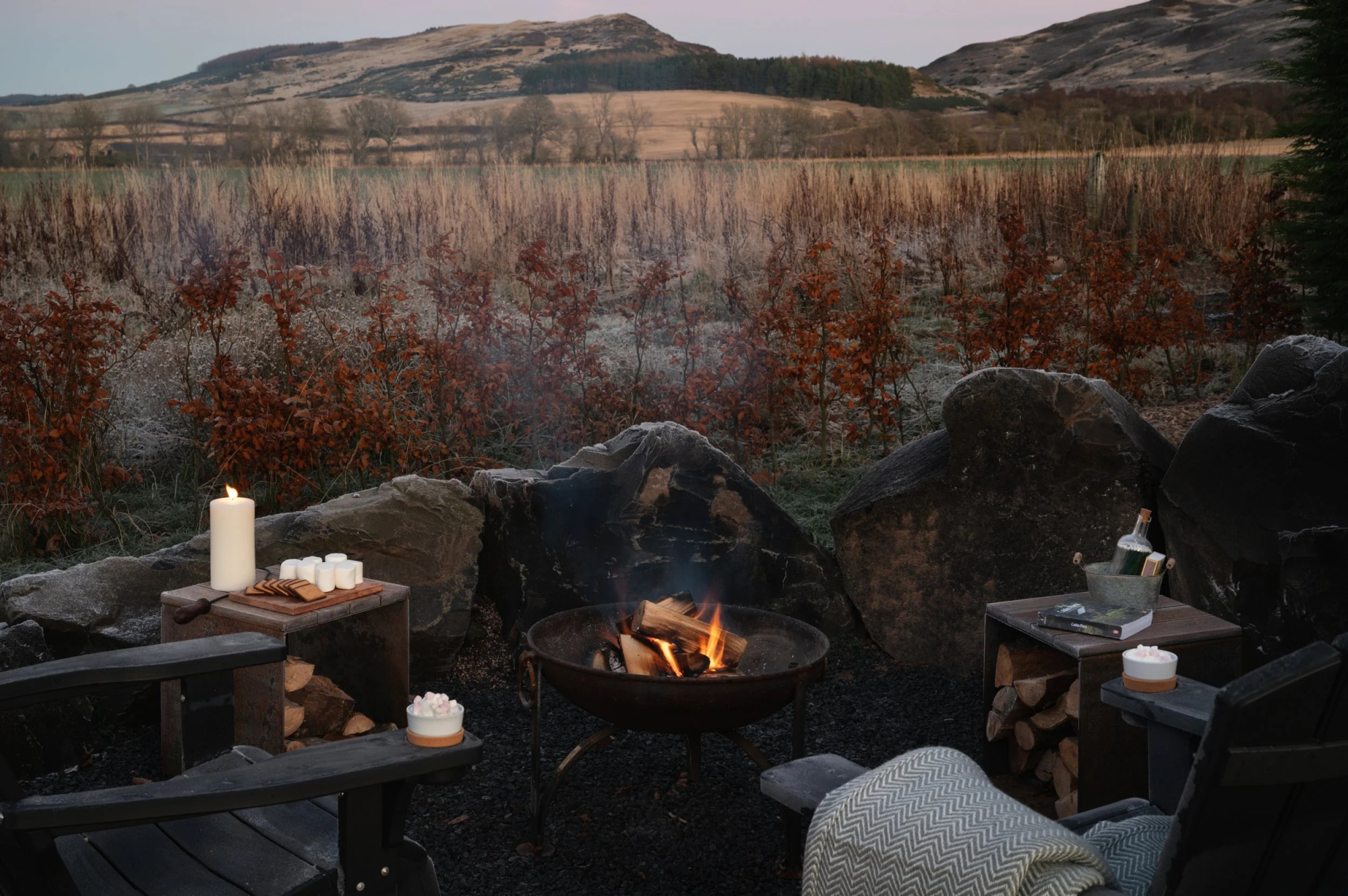 Two adirondack chairs face a lit fire bowl in a stone circle, candles and firewood on side tables, hills fading into dusk beyond frosted shrubs.