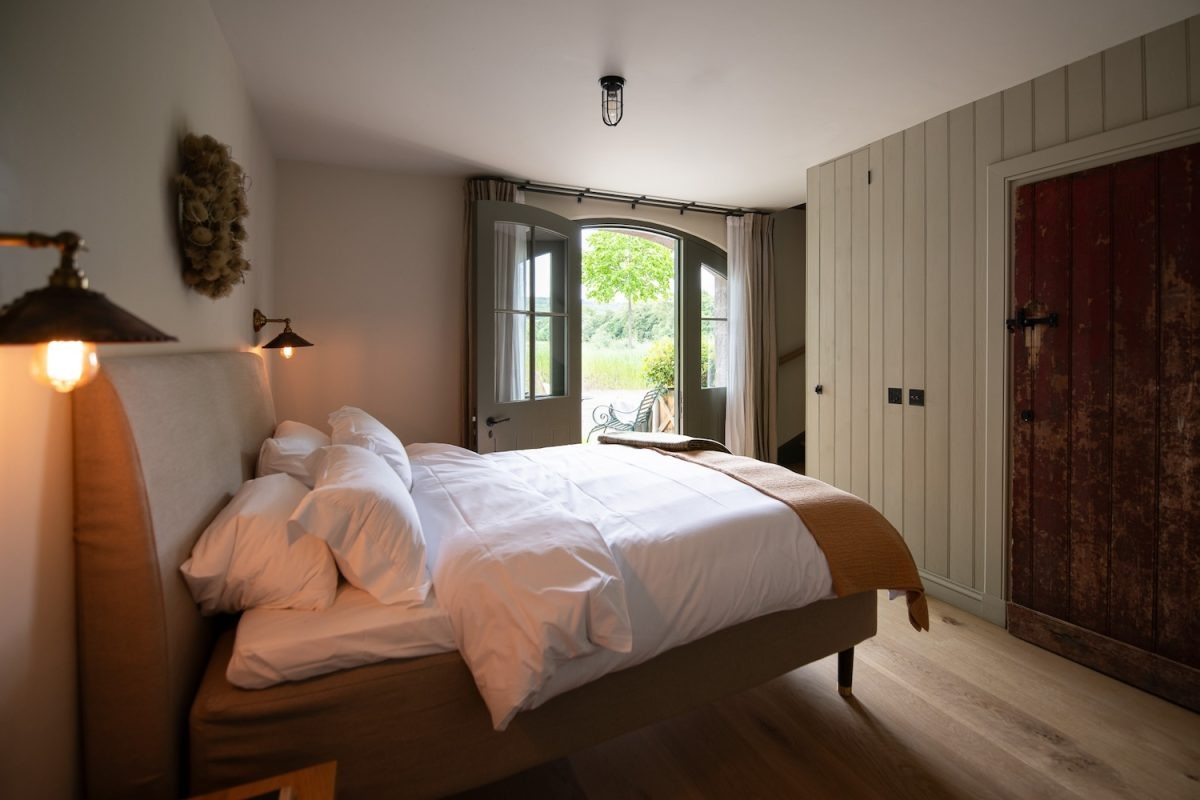 A bed with white linen and a tan throw sits before glazed doors onto a garden, a dark wardrobe and reclaimed red door to the right.
