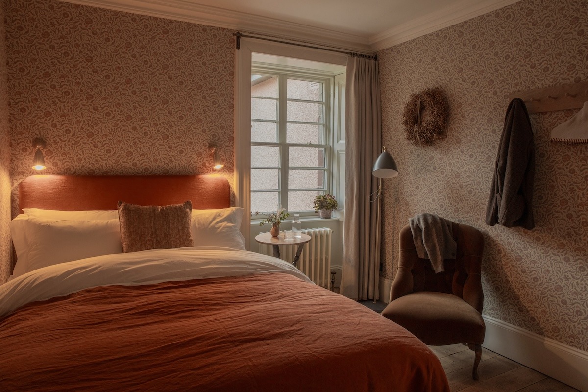 A terracotta headboard and bedspread sit against floral wallpaper, wall lamps lit either side and a wreath hanging beside a wicker chair.