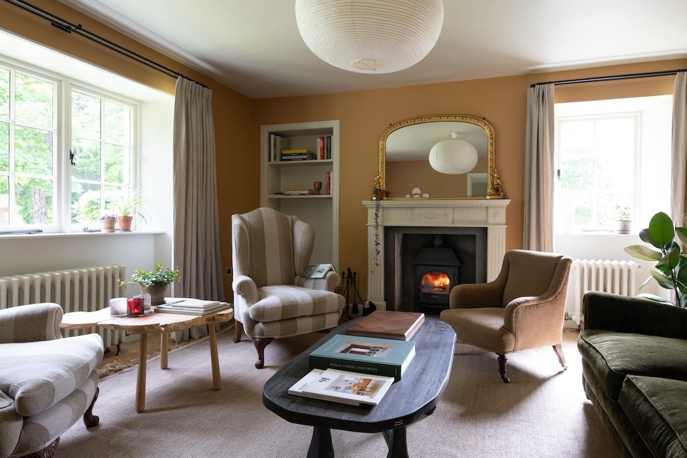 Striped and check armchairs surround a low table before a fireplace with a lit stove, a paper globe pendant light hanging from the ceiling.