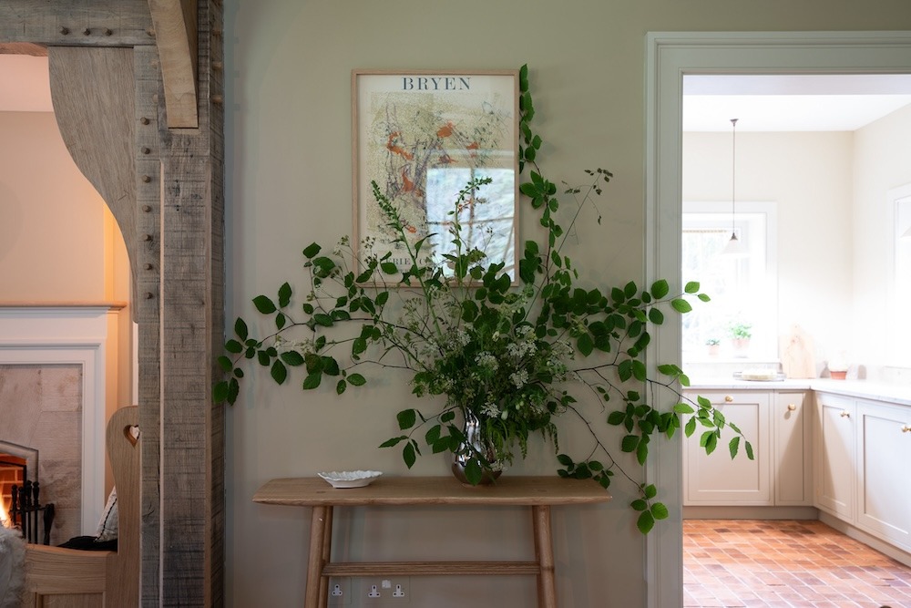 A framed print titled "Bryen" hangs above a wooden console table holding a large vase of foliage, a kitchen visible through the doorway beyond.