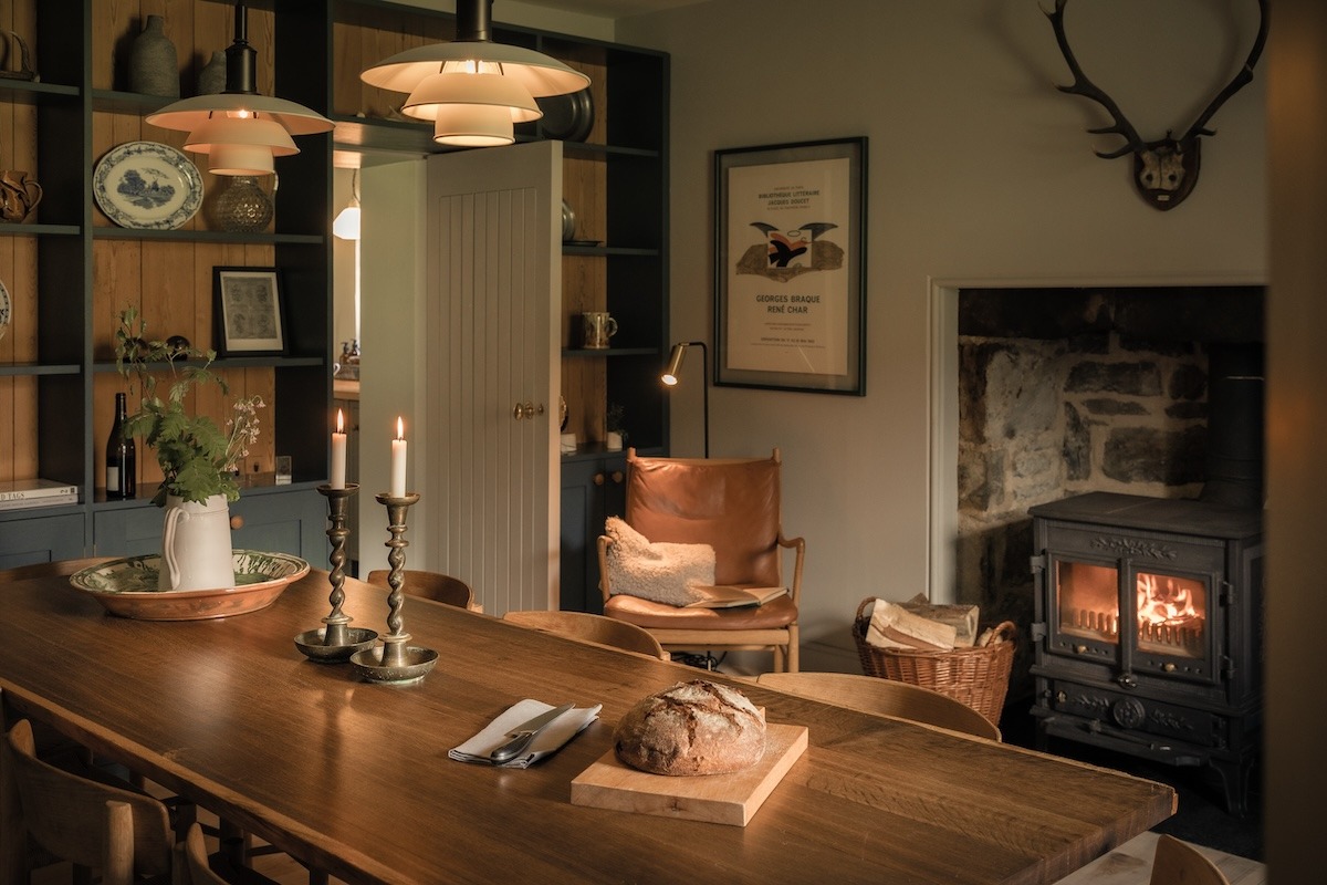 A wooden table set with a candle, bread and a jug of flowers stands before a wood burning stove, a leather armchair and framed print visible in the room behind.