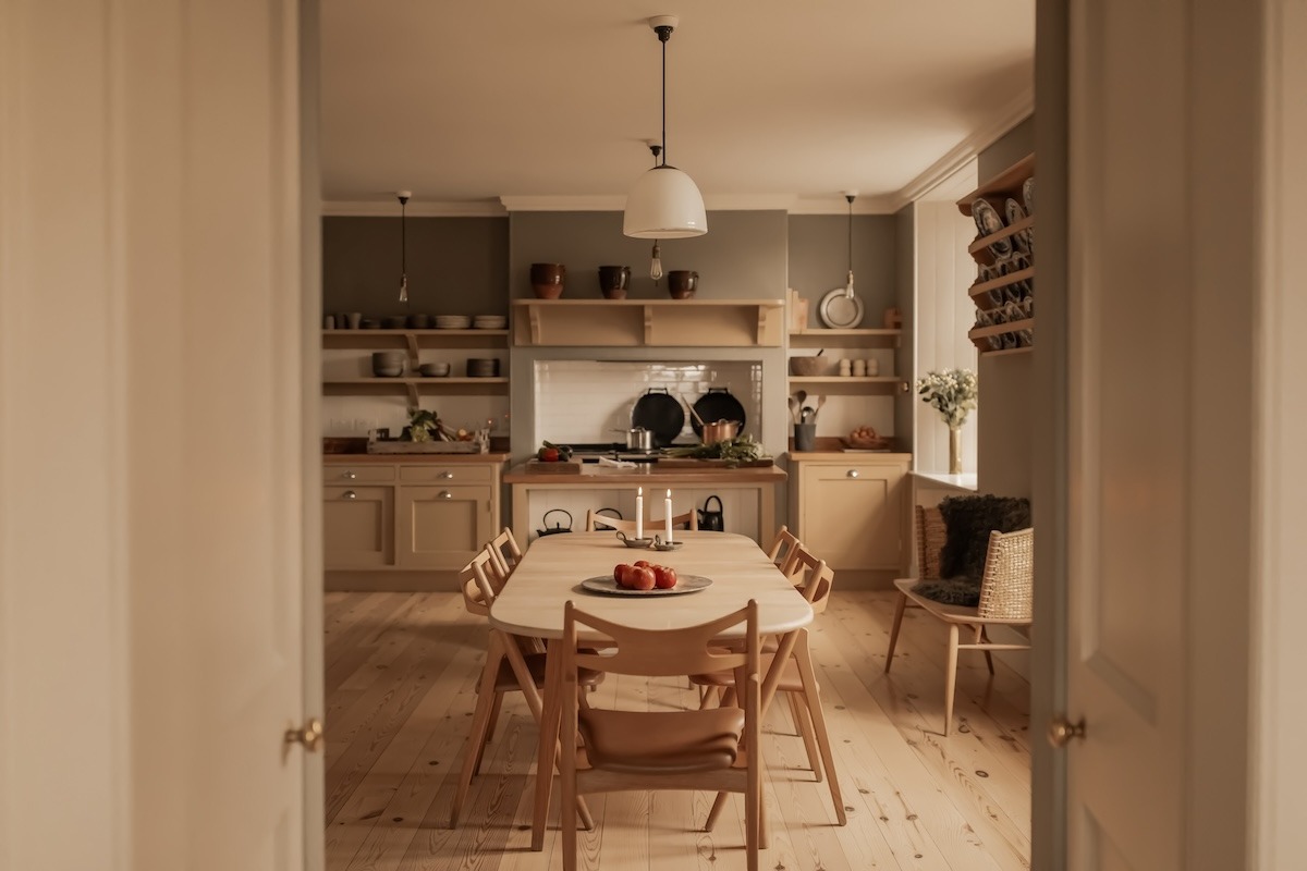 A kitchen with open shelving and a range cooker is seen through open double doors, a wooden dining table set with fruit and candles in the foreground.