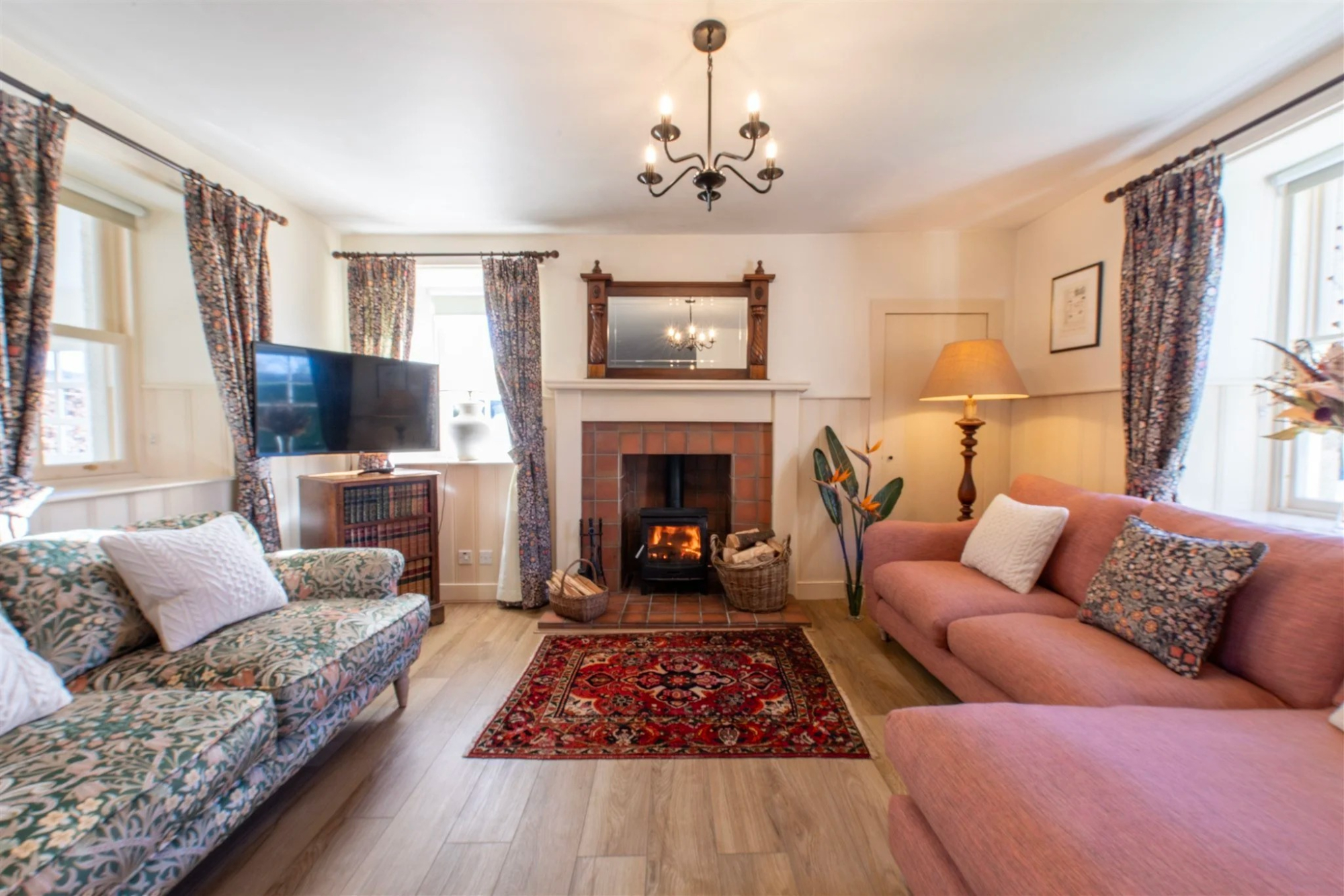 Two floral sofas face a lit fireplace under a mirror, framed pictures and a lamp on the wall to the right.