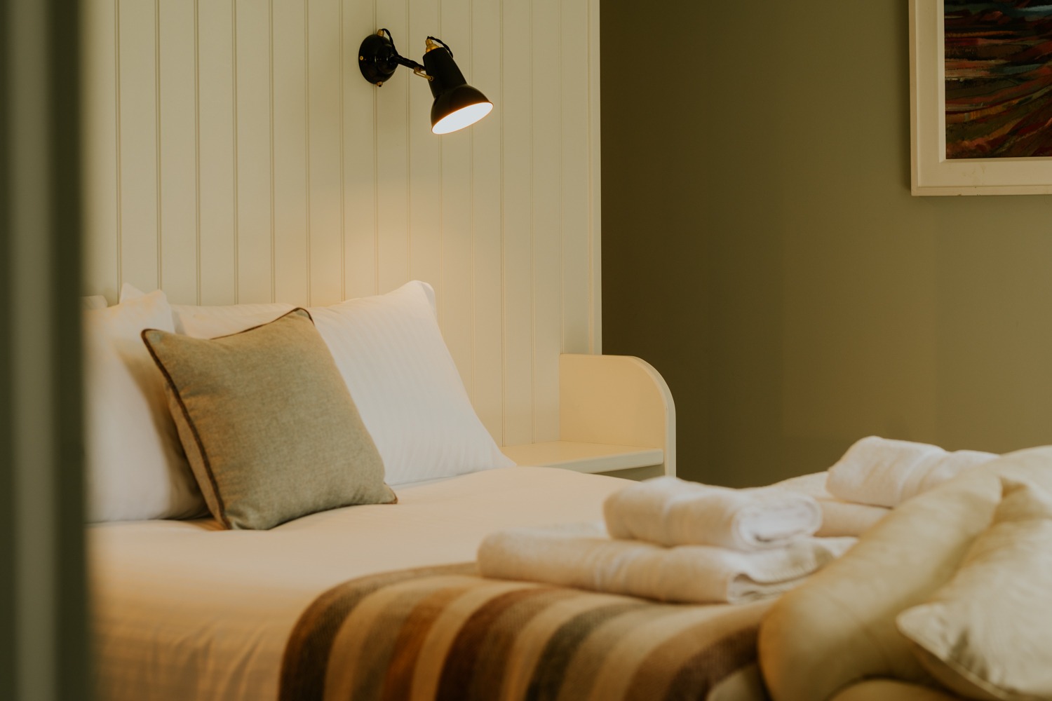 A wall-mounted lamp lights a bed with a striped throw and folded towels, a cushion and pillows against the panelled headboard.