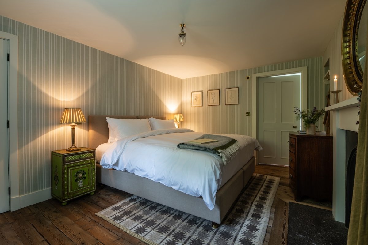 A bed with white linen sits below a gilt-framed mirror on striped wallpaper, a green bedside cabinet with a lamp to the left.