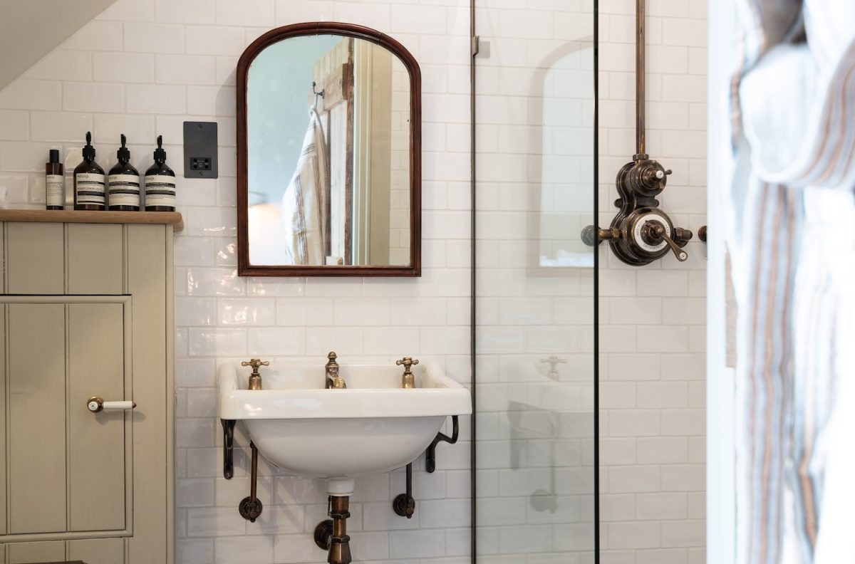 A wall-mounted pedestal basin with traditional taps stands below an arched mirror, a glass shower screen and row of amber toiletry bottles to the side.