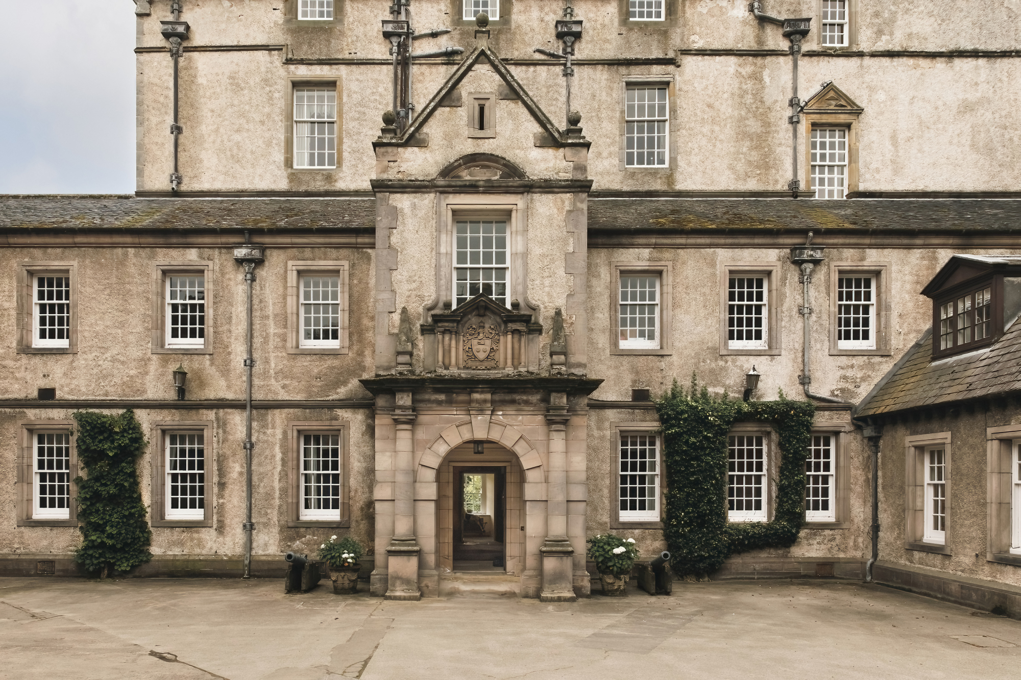A grand stone facade with an ornate carved doorway and pediment stands around a gravel courtyard, ivy climbing the walls either side.