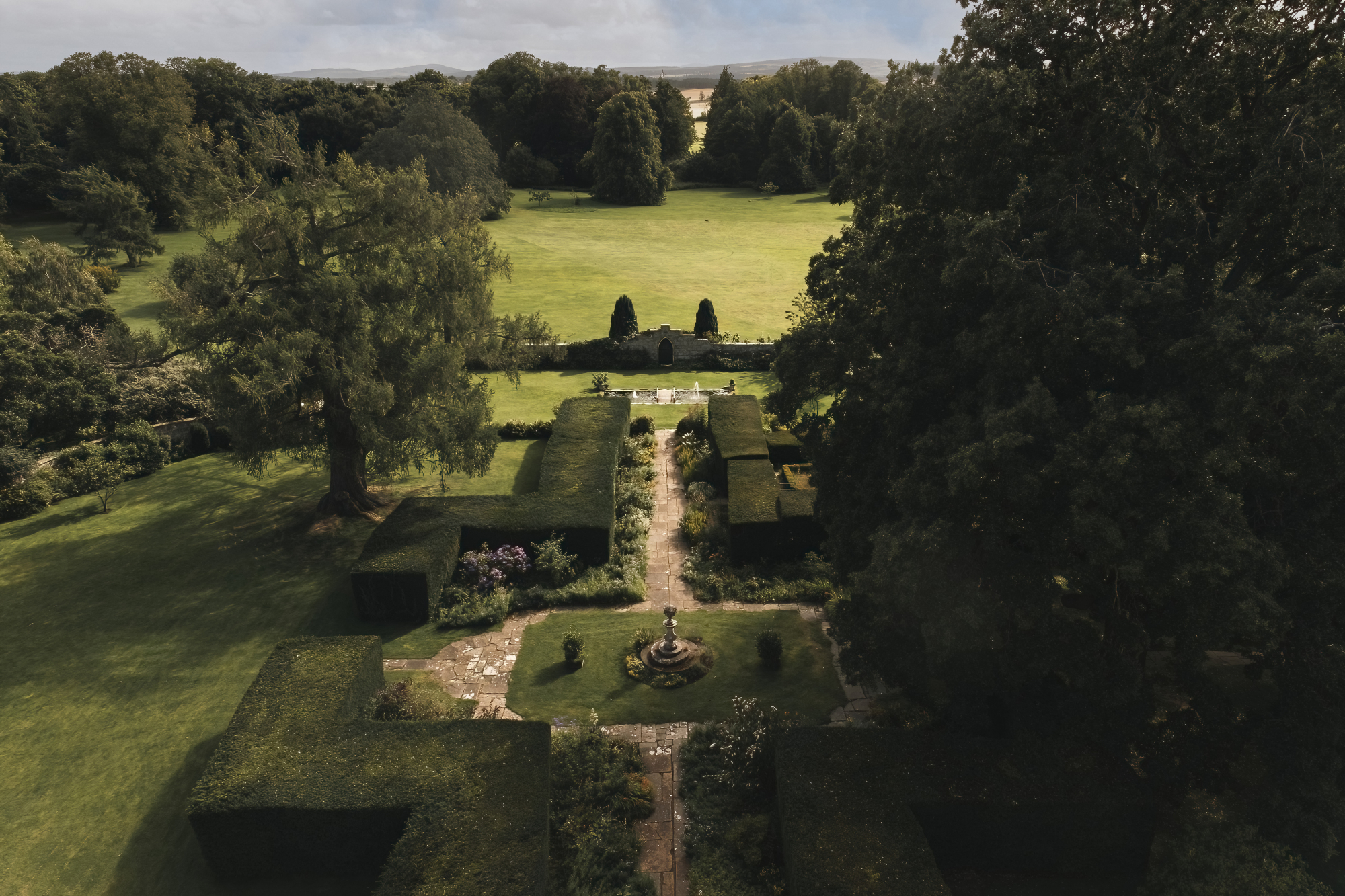 An aerial view shows a formal garden of clipped hedges and gravel paths leading to a lawn and parkland beyond.