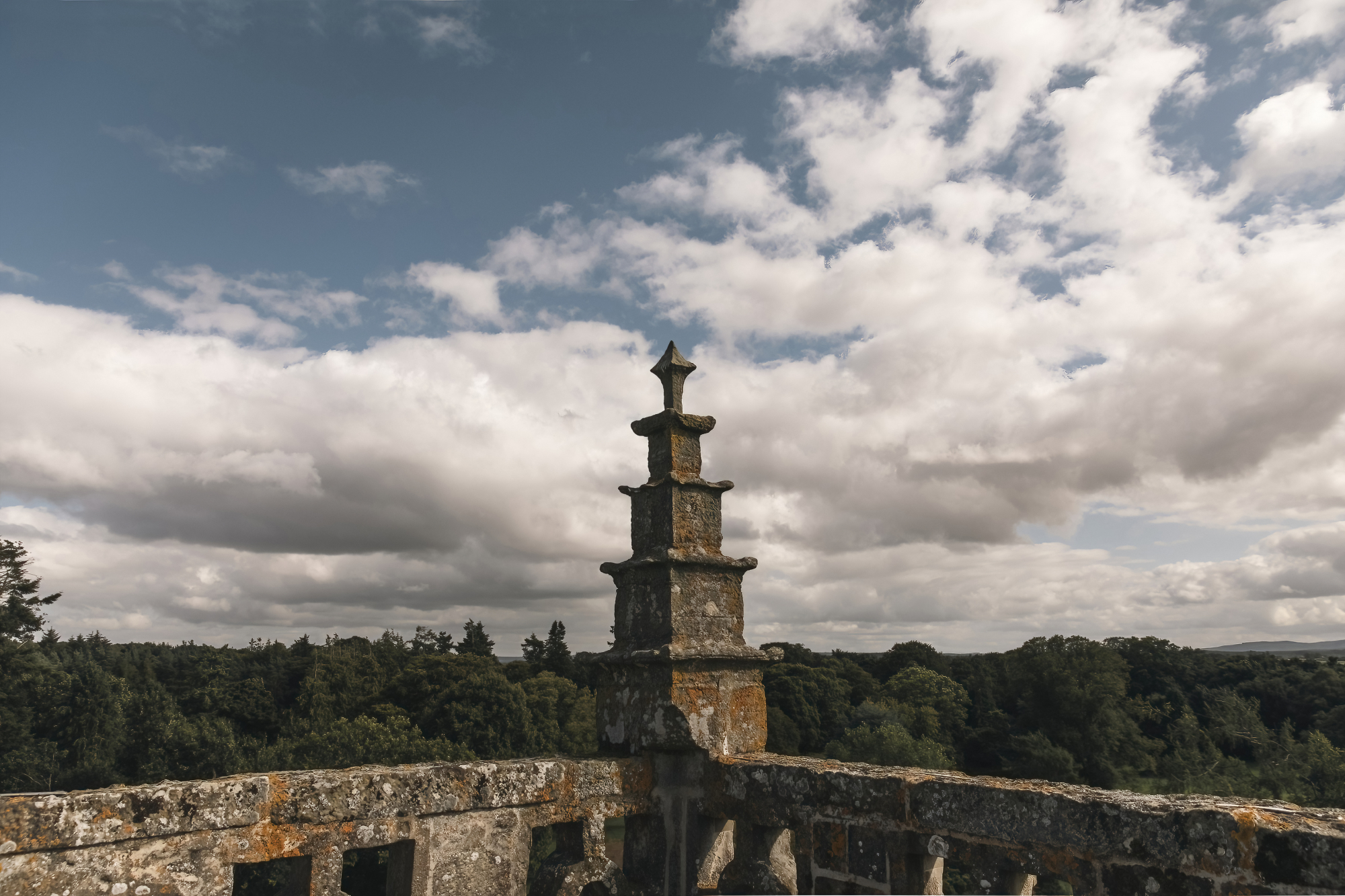 A stone finial tops a balustraded rooftop terrace, woodland stretching into the distance under a cloudy sky.