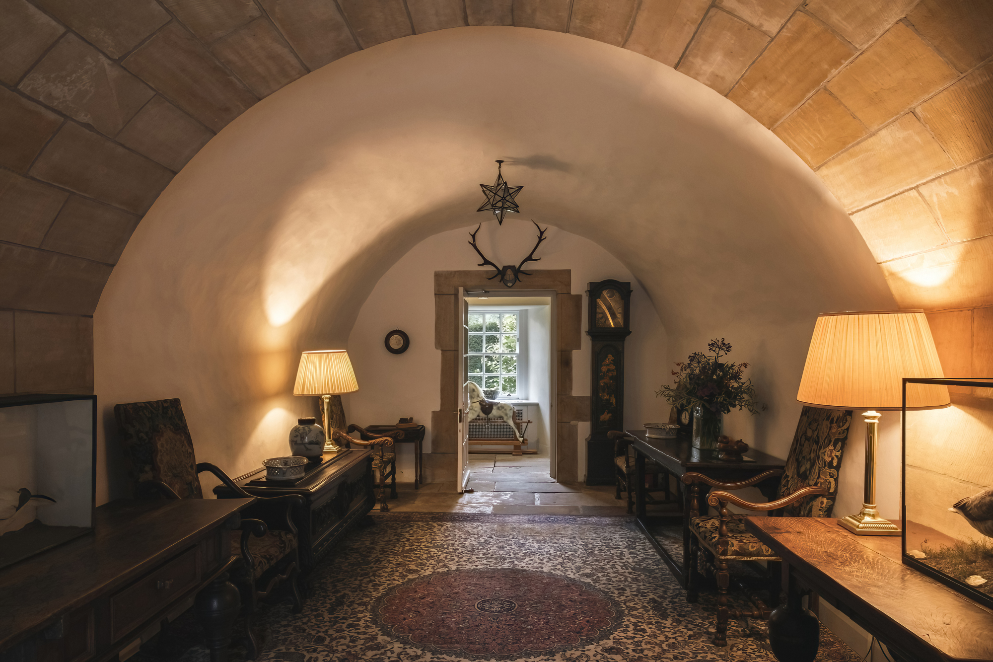 A vaulted stone hallway lit by lamps leads to an open door, antlers mounted above and a patterned rug on the tiled floor.