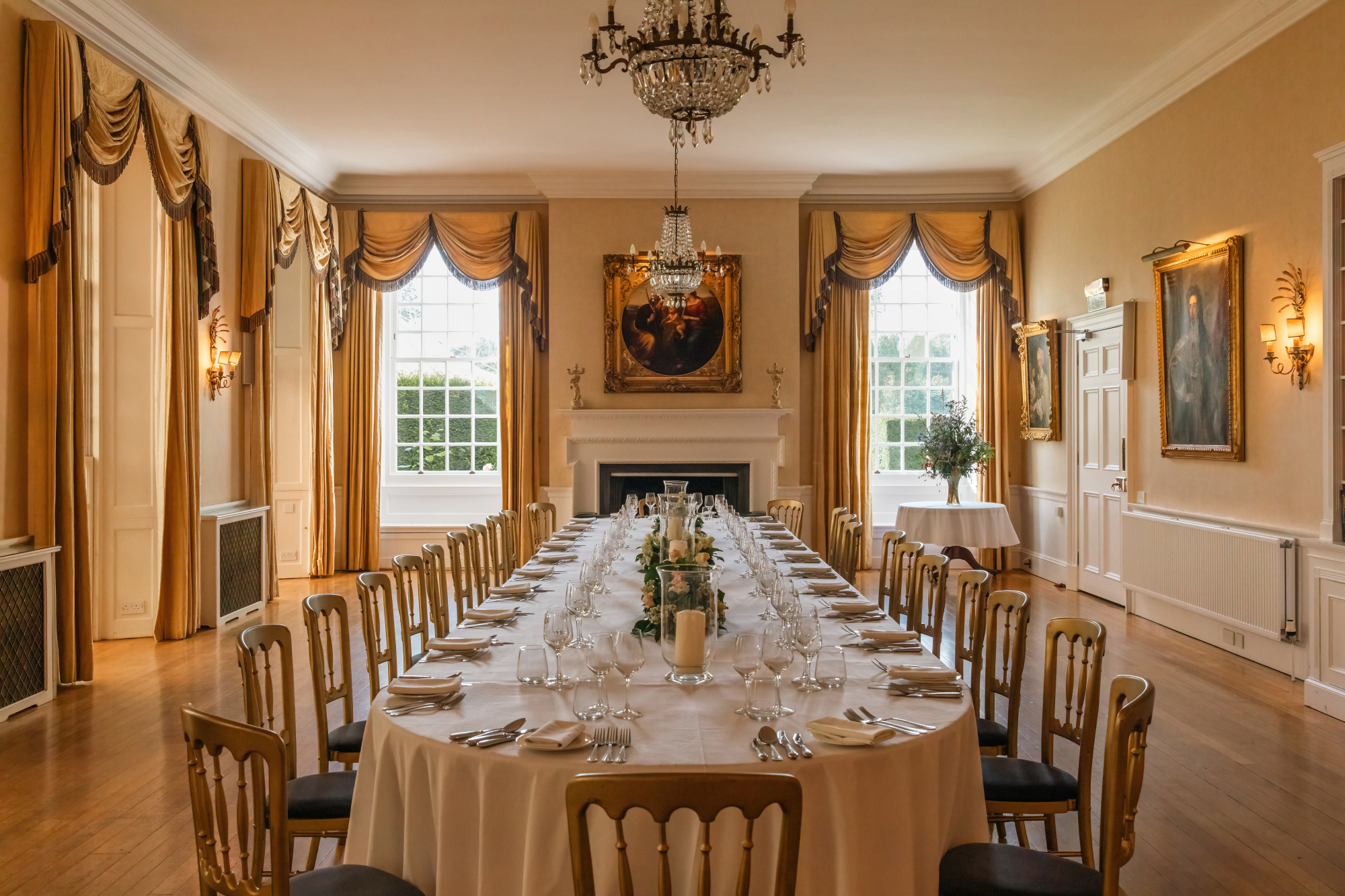 A long dining table set for a formal meal stands beneath a chandelier, gilt-framed portraits and curtained windows lining the walls.