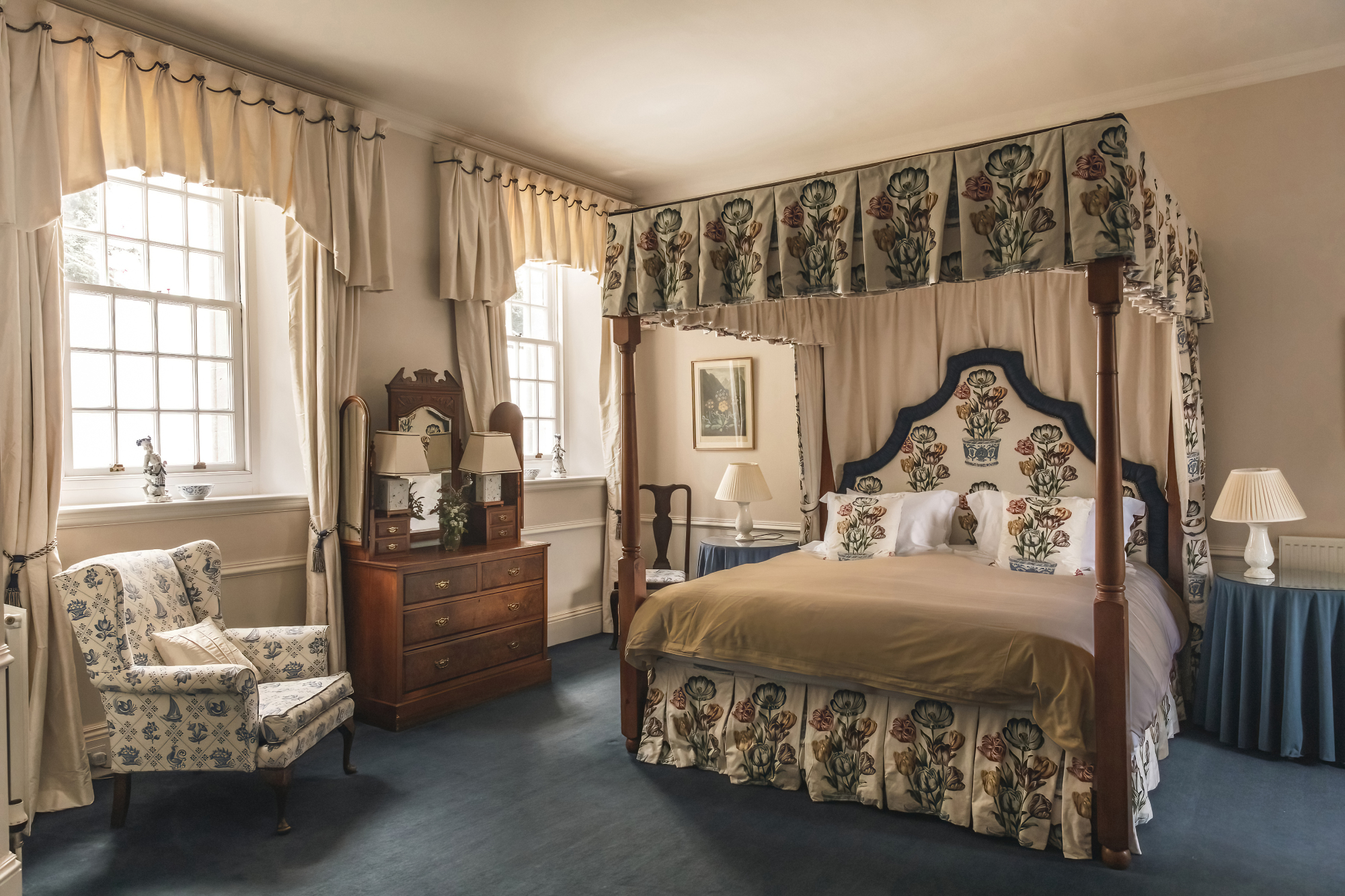 A four-poster bed with a floral canopy and matching curtains stands beside a striped armchair and dressing table, a blue throw draped over a chair to the right.