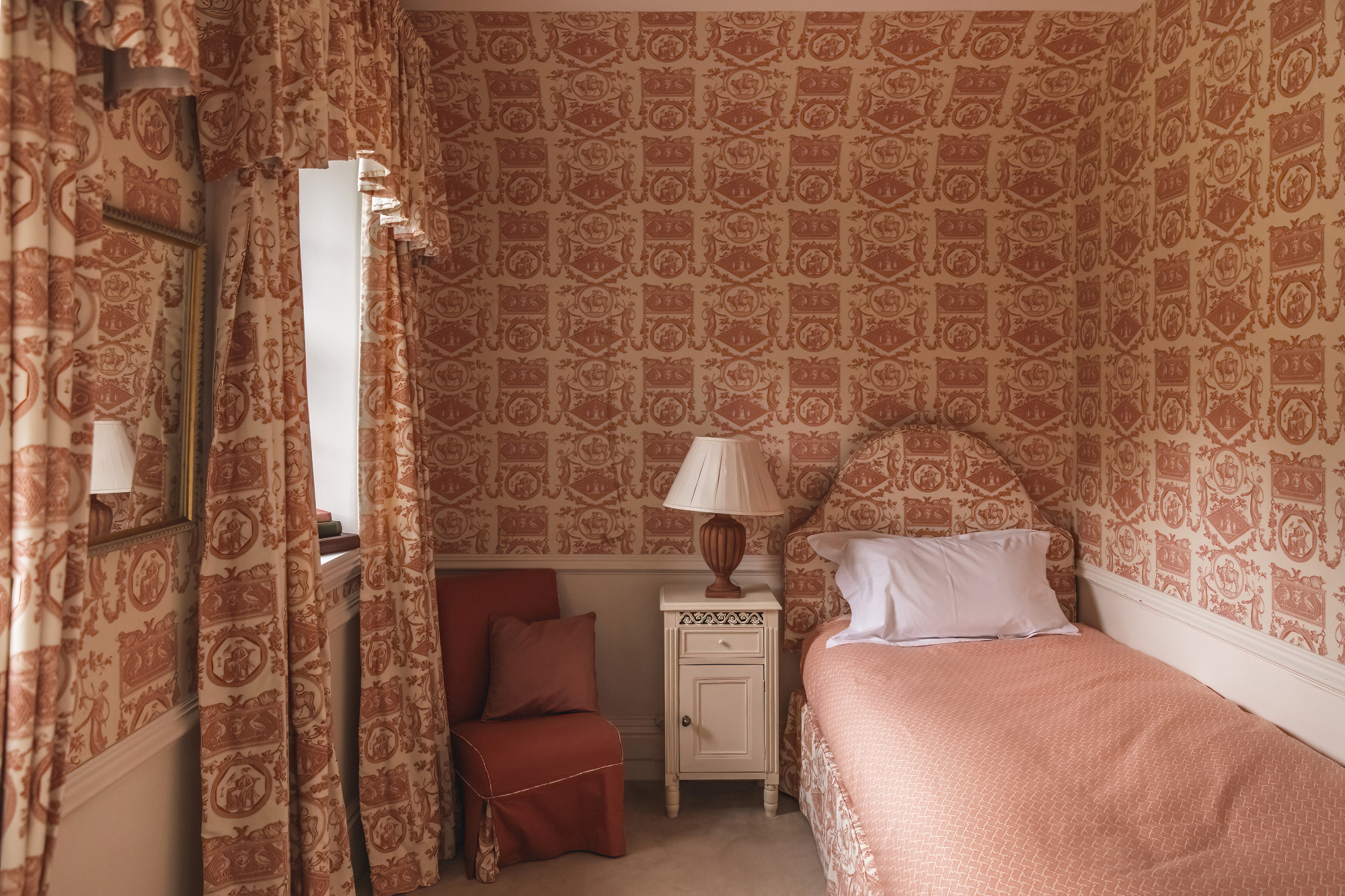 A single bed with a pink upholstered headboard sits in a room papered in matching pink toile, a white bedside cabinet and armchair completing the room.