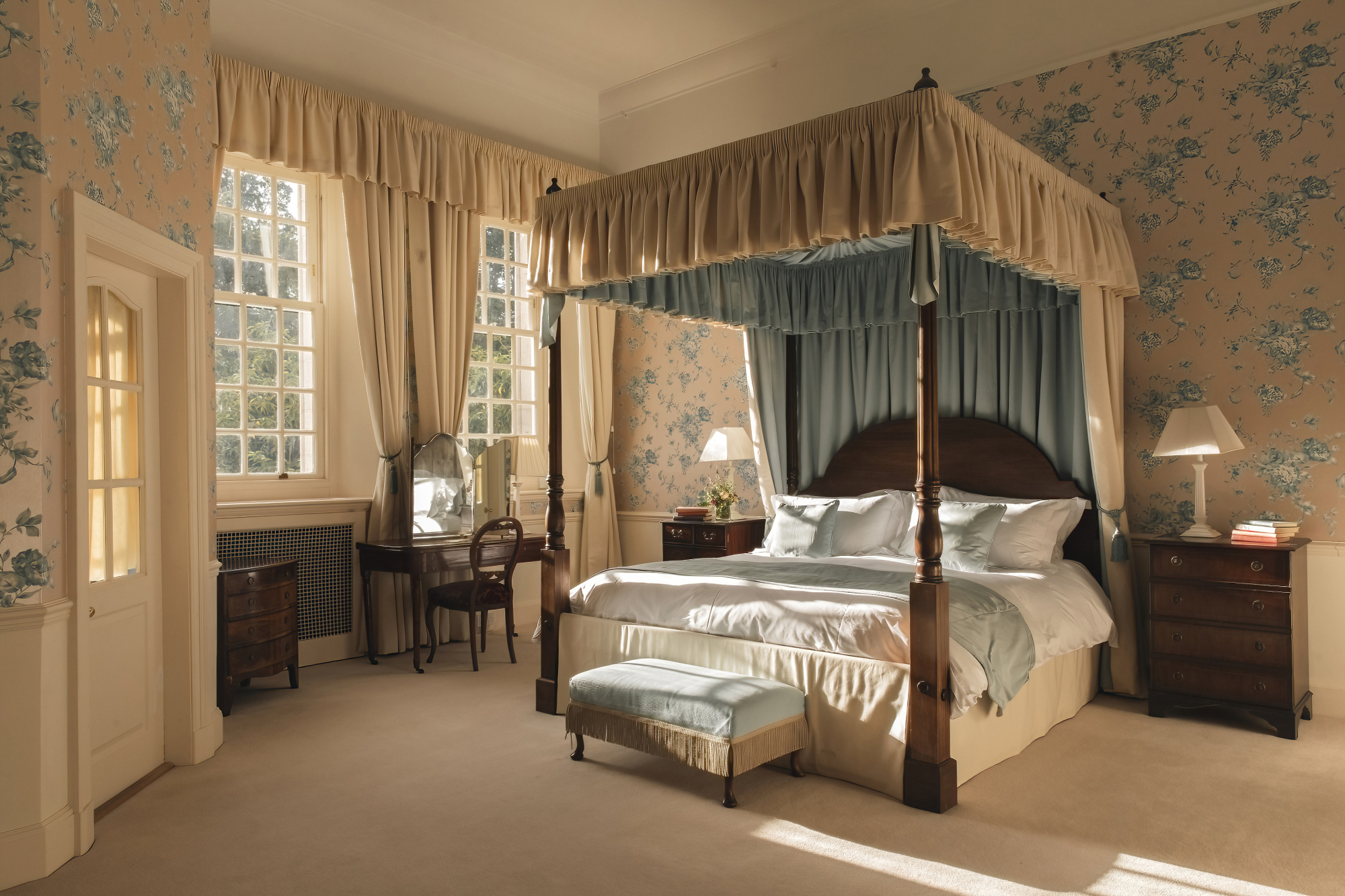 A four-poster bed with a blue canopy and floral drapes stands in a room papered with matching blue florals, a dressing table and bench at its foot.