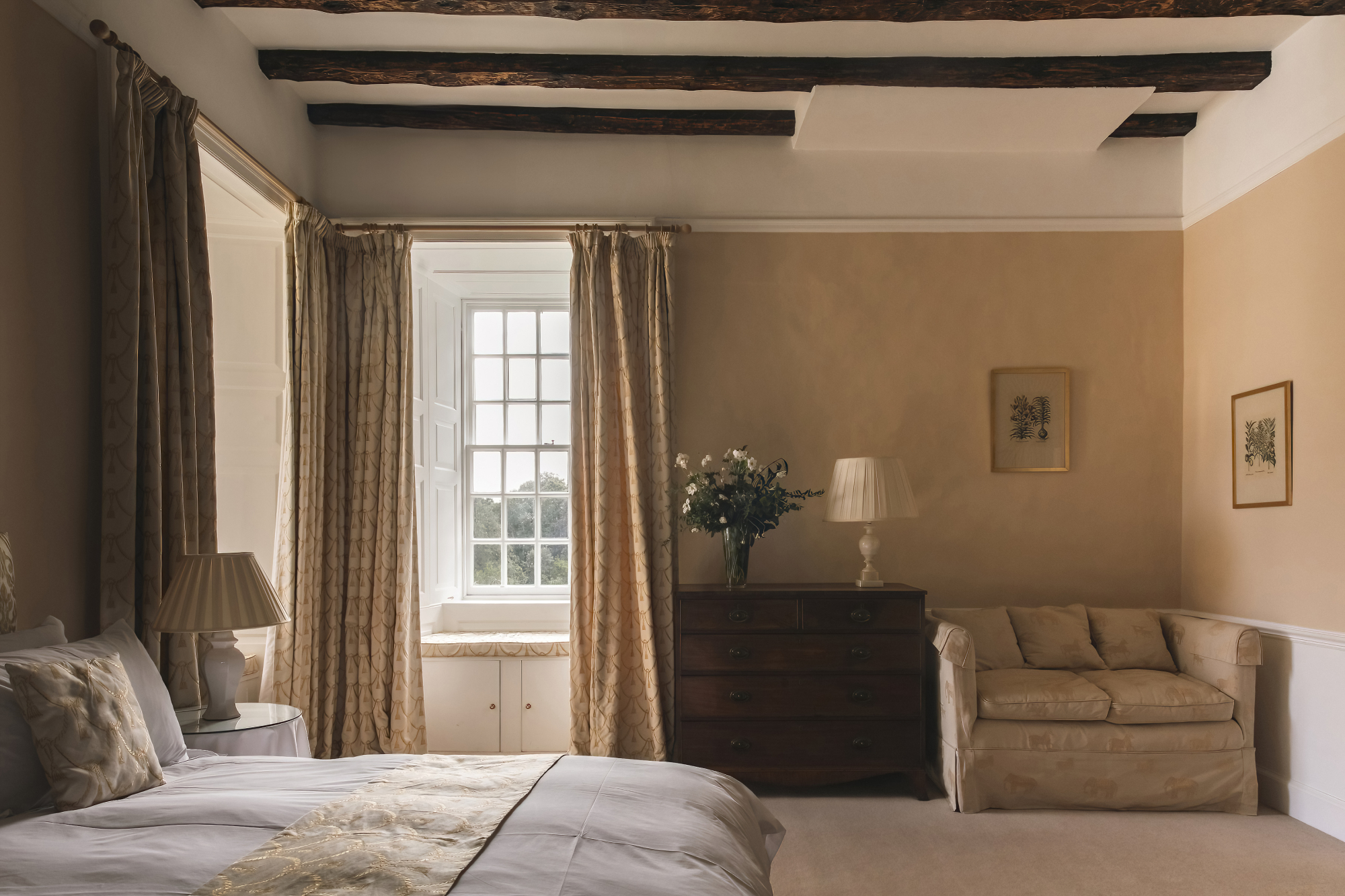 A bed with white linen sits below exposed ceiling beams, a chest of drawers with a vase of flowers and an armchair by the window.