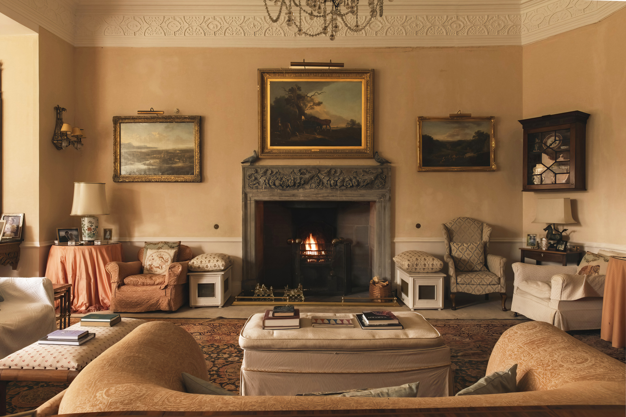 Cream sofas and armchairs surround an ottoman before a lit fireplace, landscape paintings hung above the mantel and on the walls either side.