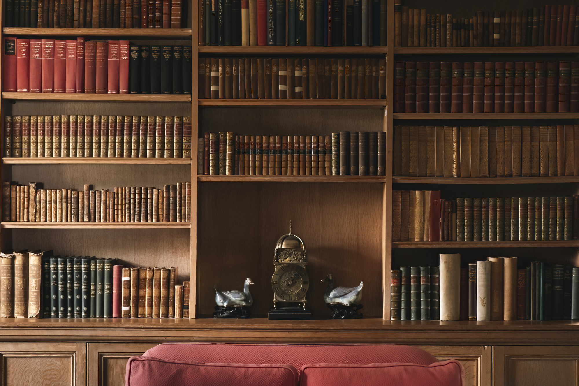 A carriage clock flanked by two carved duck ornaments sits on a shelf between floor-to-ceiling bookcases, a red sofa in the foreground.