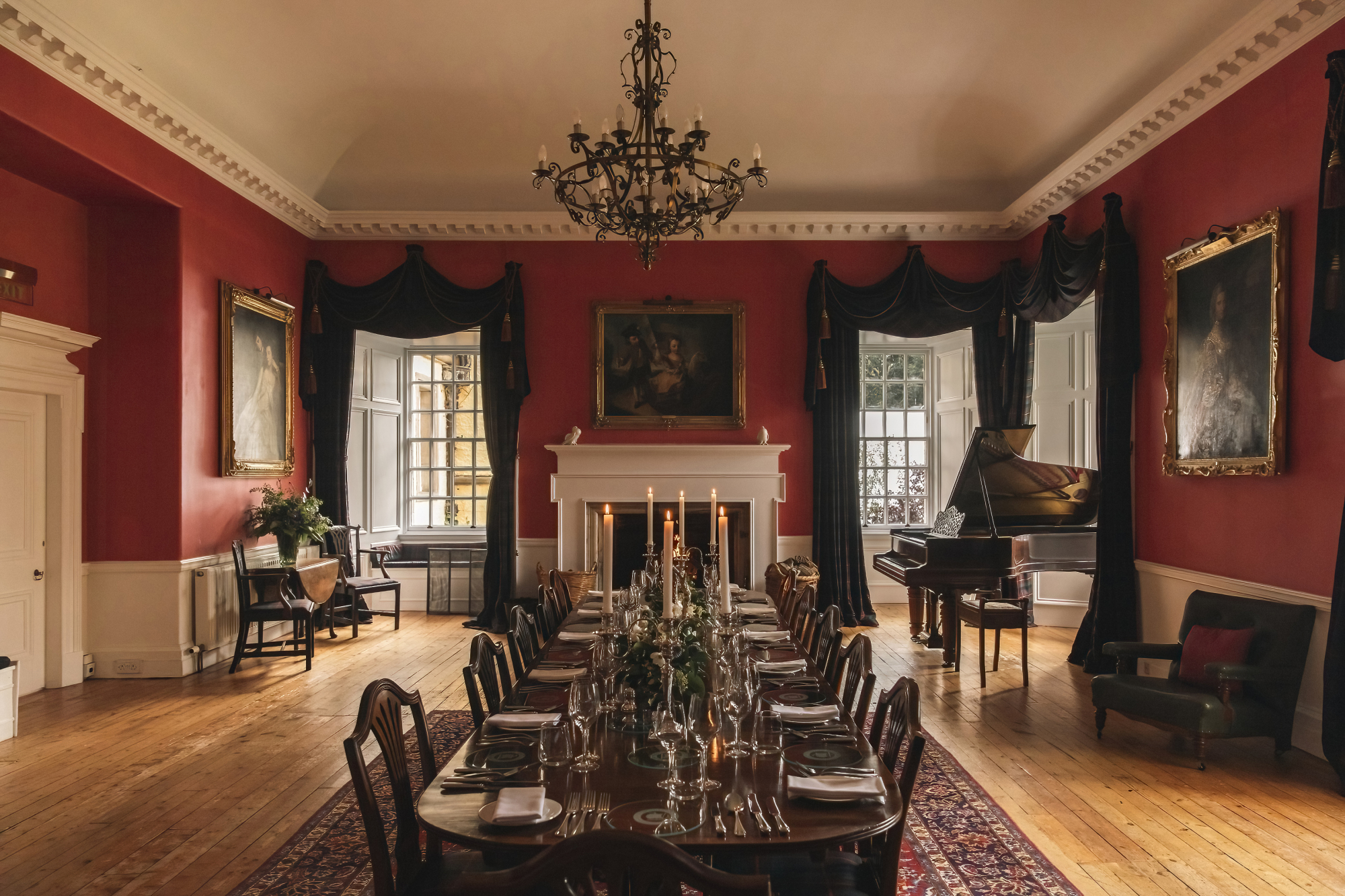 A long dining table set with candles and flowers stands before a fireplace in a red-walled room, a grand piano visible through the archway beyond.