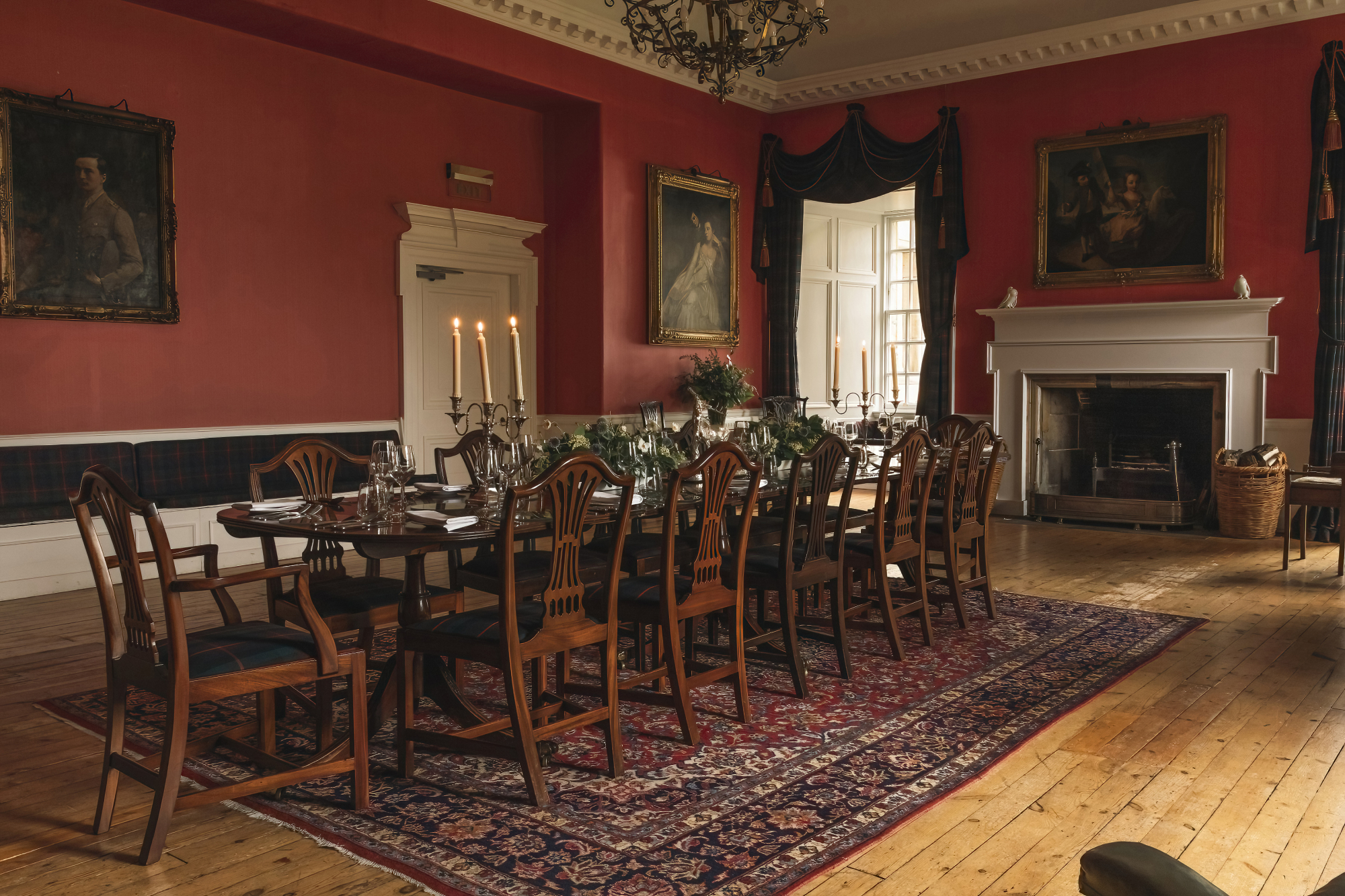 A long dining table set for a formal meal with candelabra and flowers stands on a patterned rug before a red-walled room hung with portraits, a fireplace to the right.