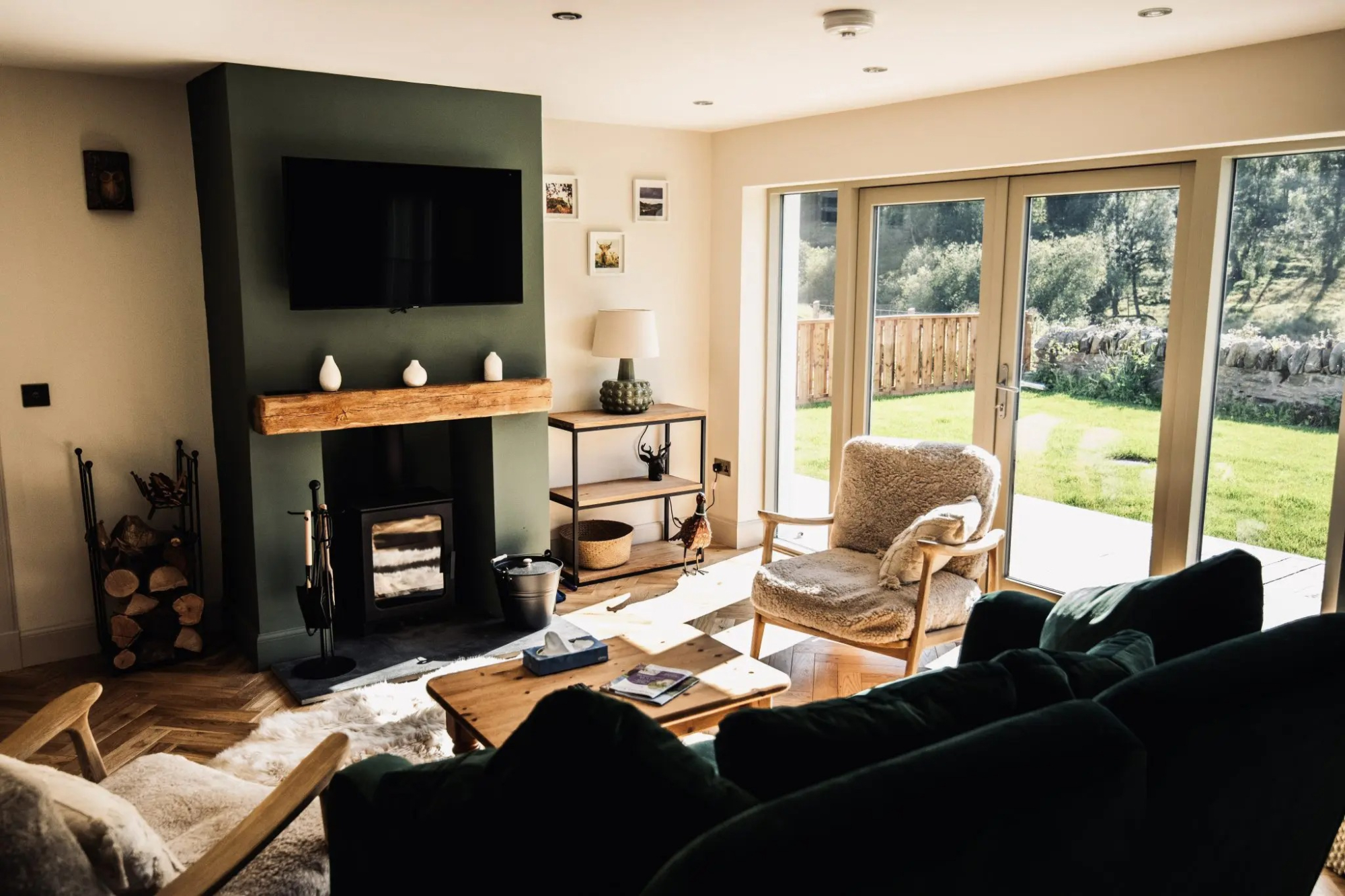 A wood burning stove sits below a mounted television on a green chimney breast, log baskets to the side and a green sofa in the foreground before glazed doors onto a garden.