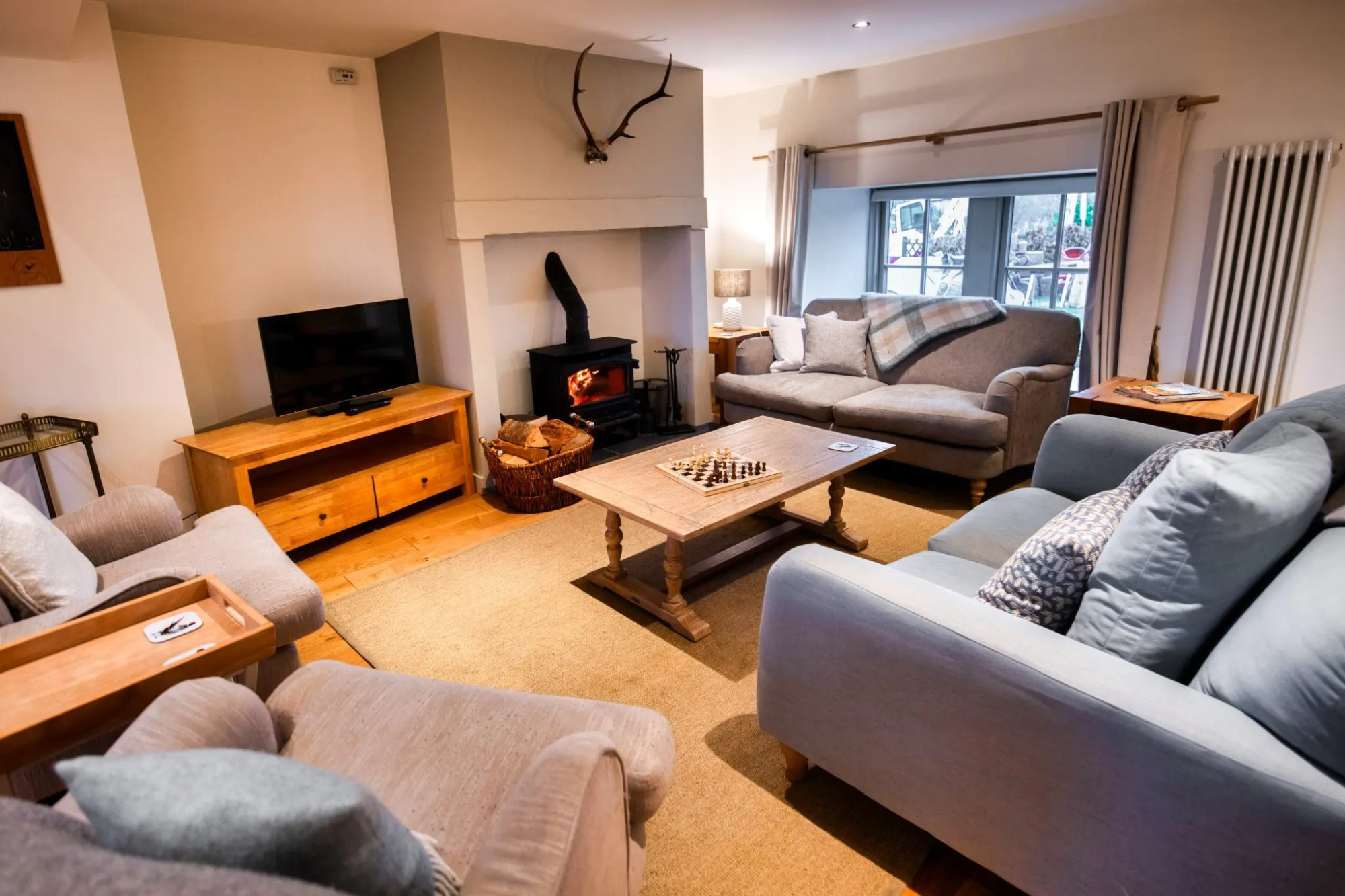 A wood burning stove sits in a stone fireplace below mounted antlers, sofas and armchairs arranged around a chess table on a patterned rug.