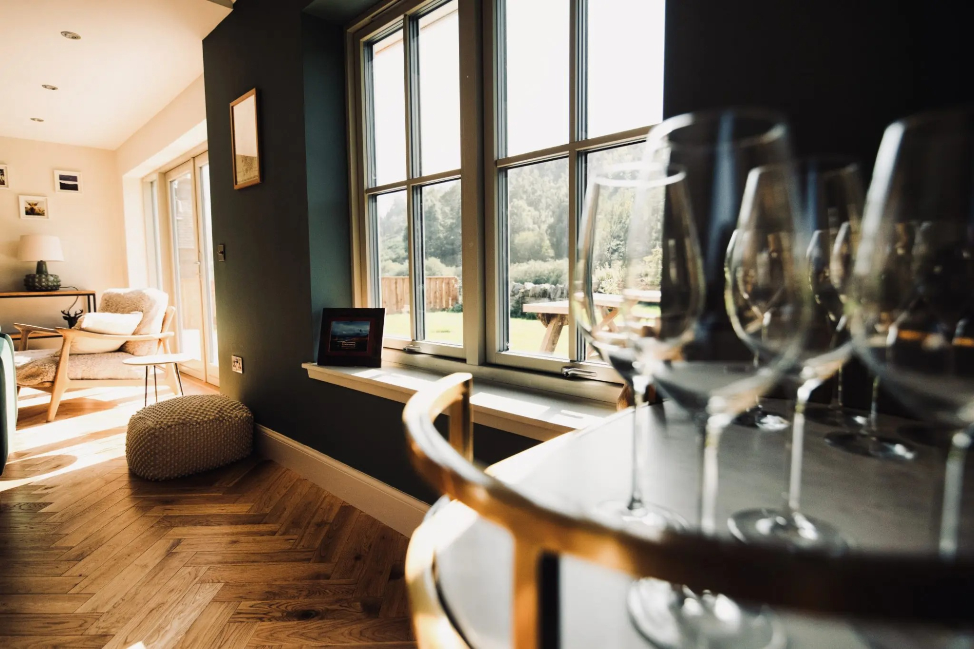 Wine glasses stand on a gold-rimmed table beside a window overlooking a garden, a sitting area visible beyond a low blue wall.