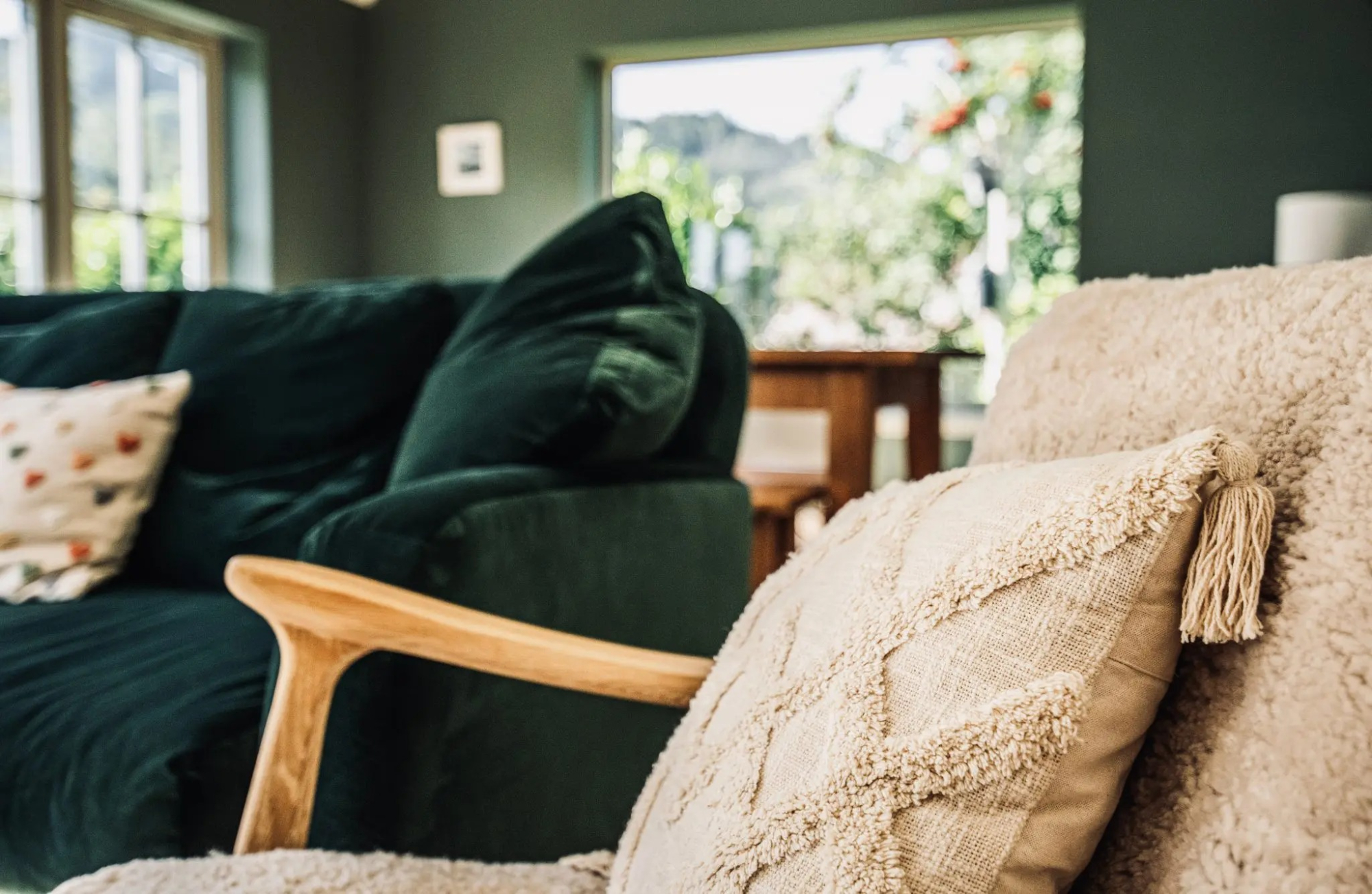 A cream tasselled cushion rests against a bouclé armchair beside a dark green velvet sofa, a window framing hills behind.