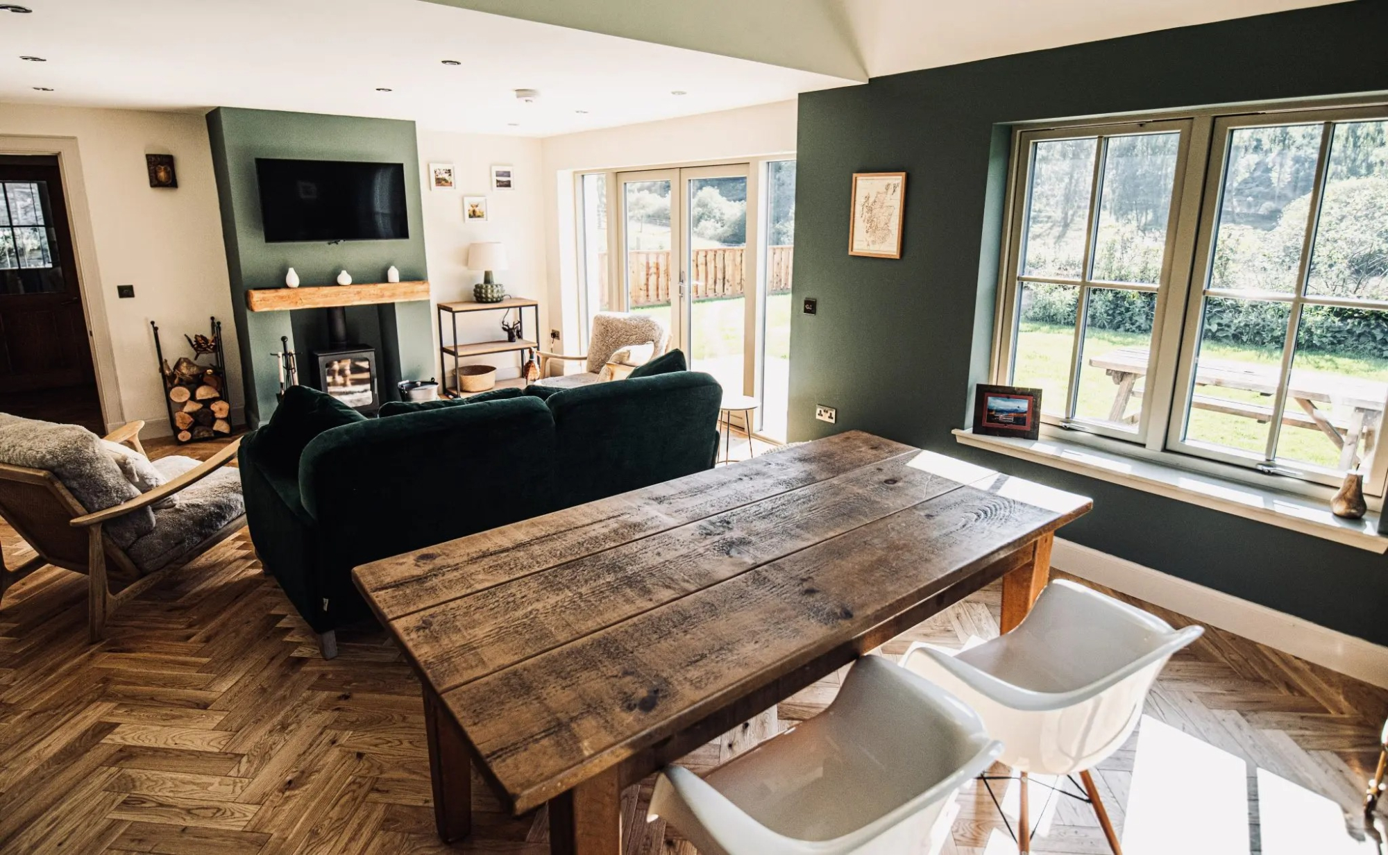 A wooden dining table with white chairs stands before a green wall and window, a living area with a sofa and stove visible behind.