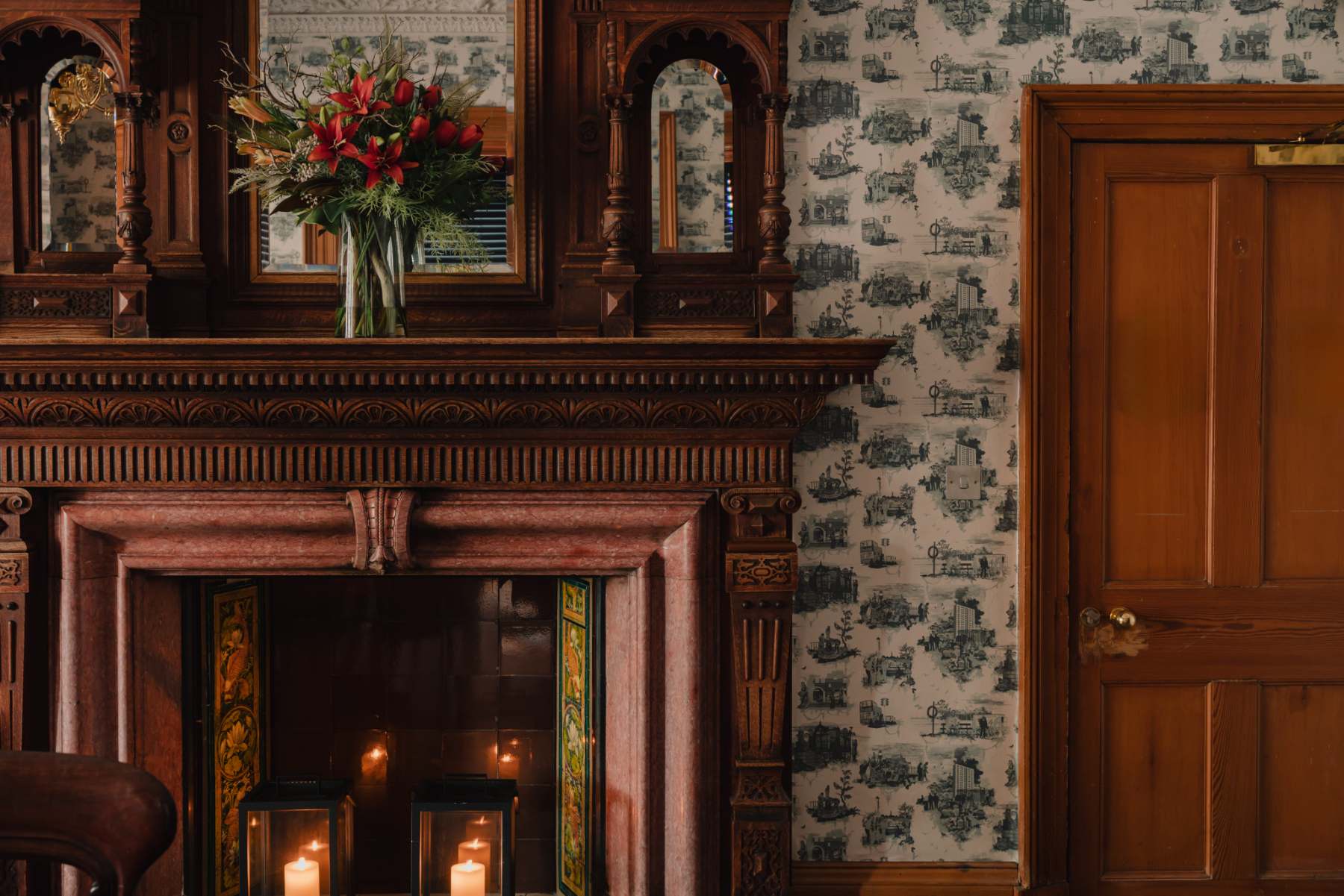 An ornate carved wooden fireplace mantel holds a vase of red flowers reflected in the mirror above, patterned wallpaper and a panelled door to the side.