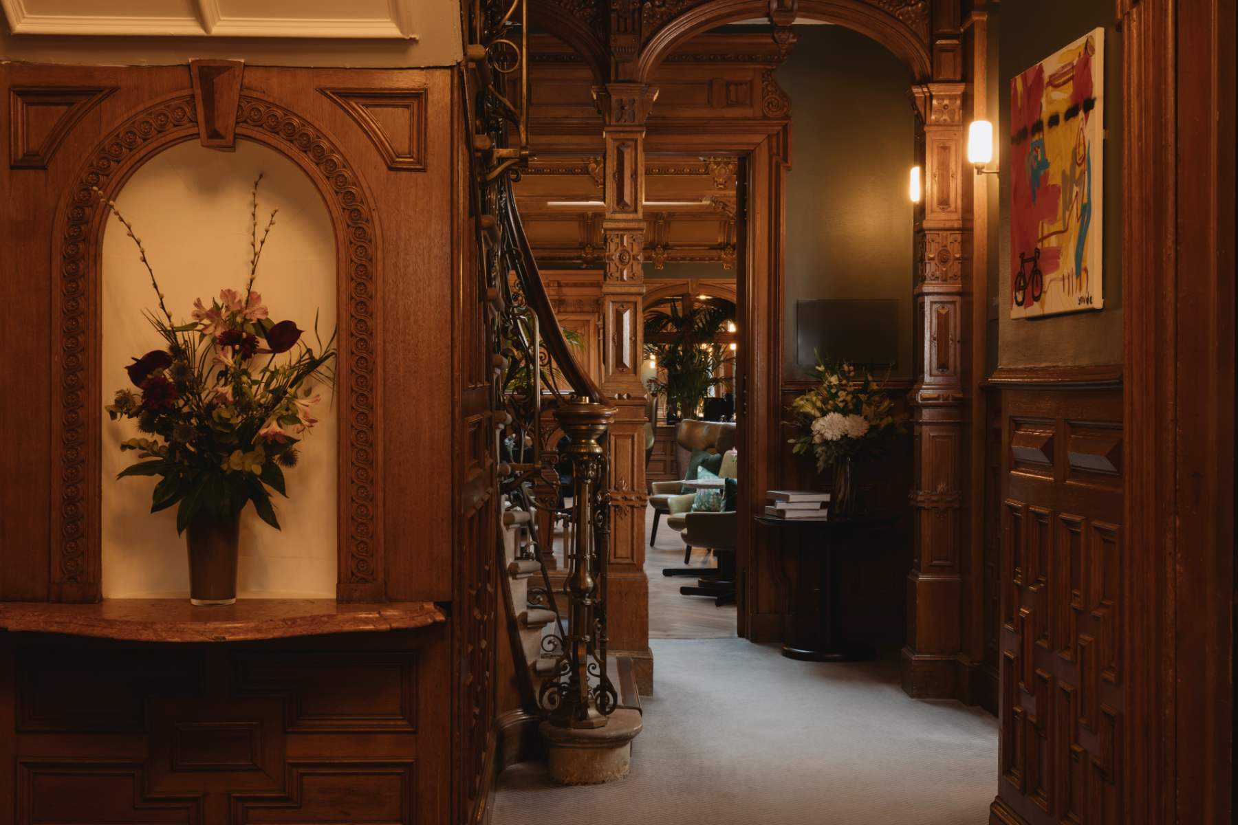 A vase of flowers stands in a wood-panelled alcove beside an ornate staircase, a colourful abstract painting on the wall.