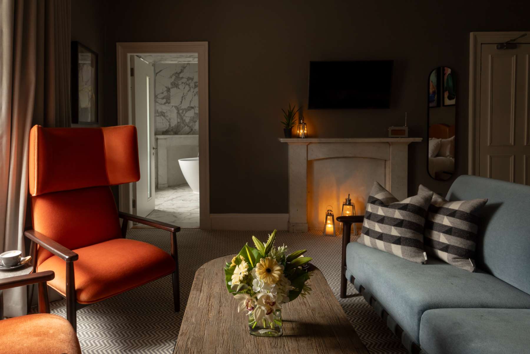 An orange wingback chair and a blue sofa with striped cushions sit around a coffee table of flowers, a lit fireplace and a bathroom visible through an open door.