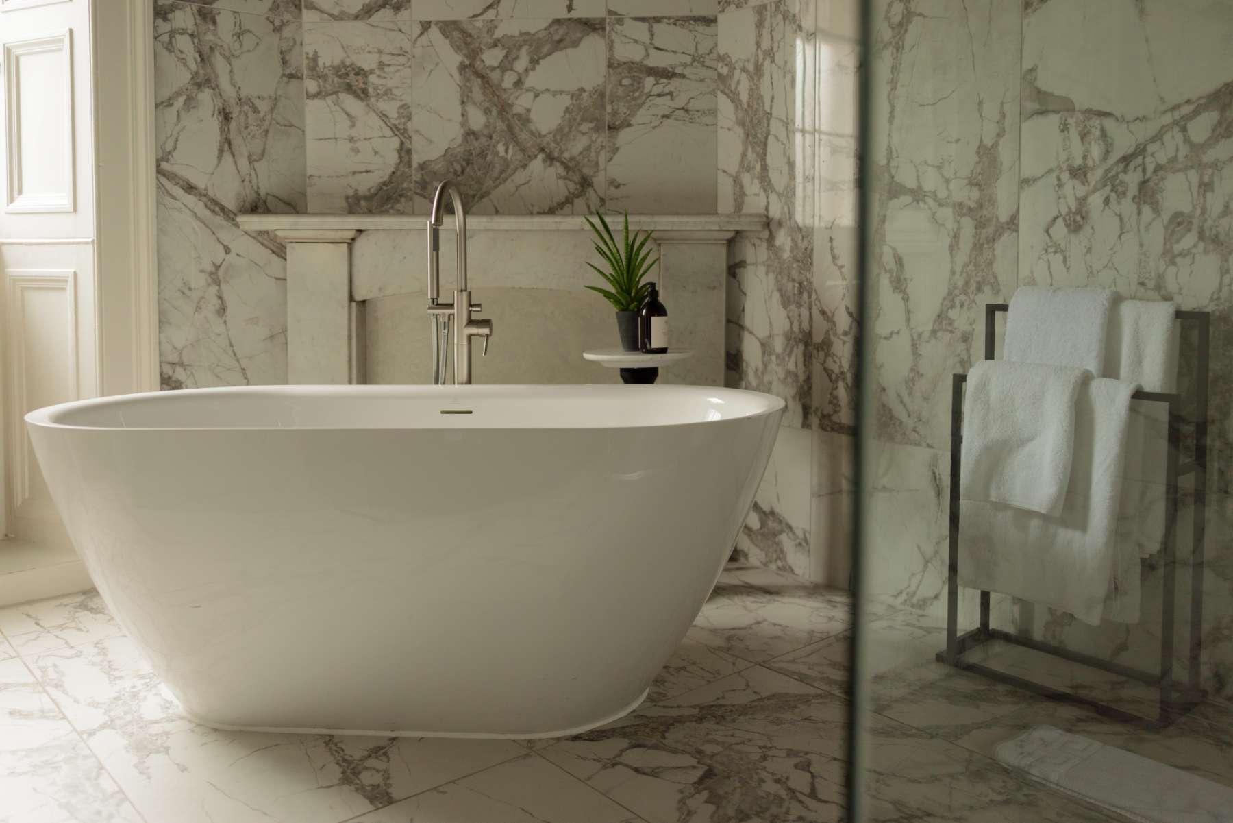 A freestanding marble bath stands beneath a chrome tap and a small potted plant, a towel rack against the marble wall behind.
