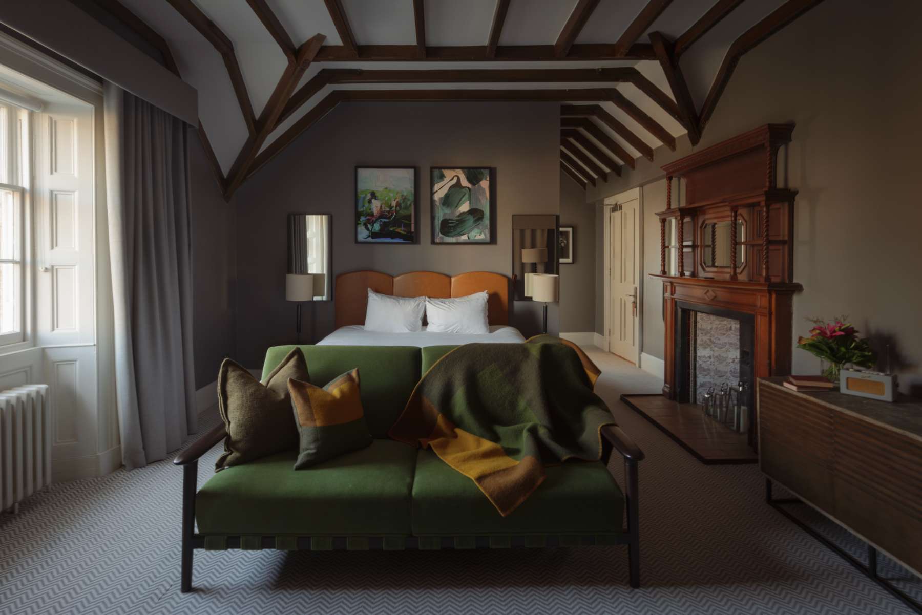 A wooden bed with an orange throw and cushions sits before a green sofa and an antique fireplace, framed art on the dark walls.