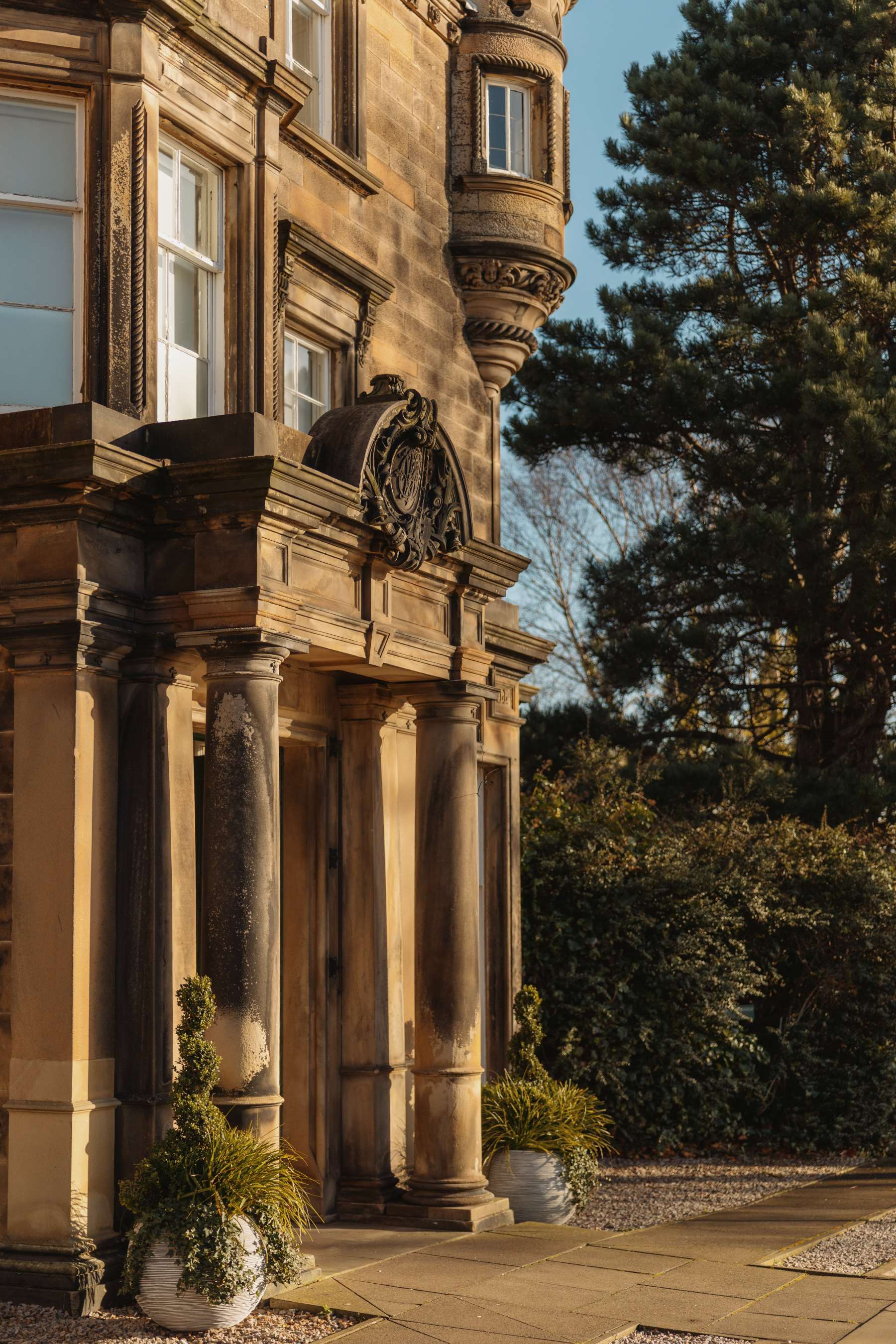 A sandstone building facade has a columned entrance porch beneath a carved stone crest, potted plants either side of the doors.